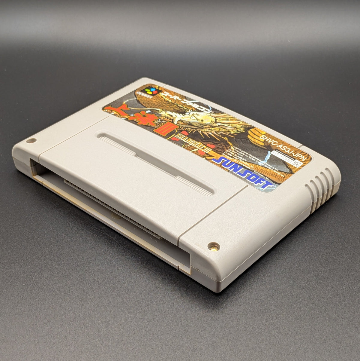 Shanghai 3 video game cartridge for Nintendo's Super Famicom (SFC) Entertainment System, shown laying on its back with the label facing upwards. Showing the right hand side and bottom slot. It is displayed on a dark grey background.