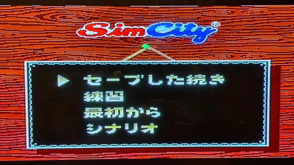 The game title screen for Sim City on the Super Famicom (SFC), with Japanese text options on a black menu box, set against a red wood-grain background with the SimCity logo at the top. This is to show the game is tested and working.