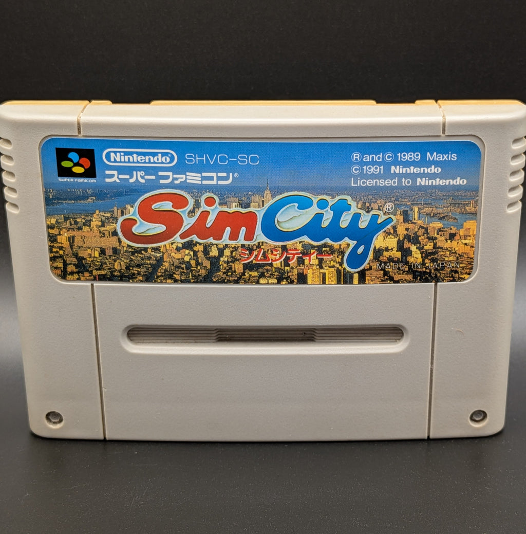A close up of Sim City video game cartridge for Nintendo's Super Famicom (SFC) Entertainment System, shown standing up and facing forwards on a dark grey background.