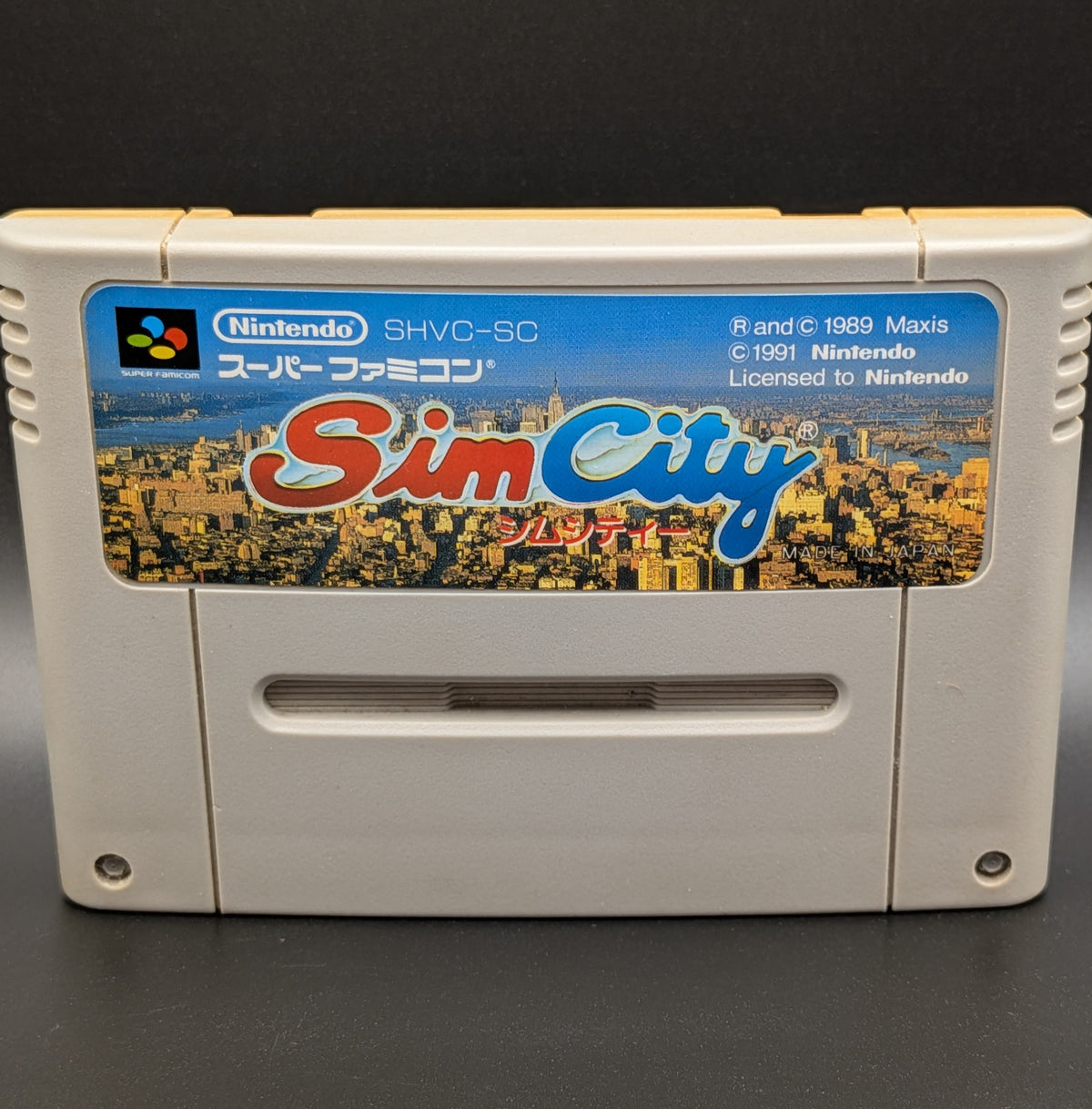 A close up of Sim City video game cartridge for Nintendo's Super Famicom (SFC) Entertainment System, shown standing up and facing forwards on a dark grey background.