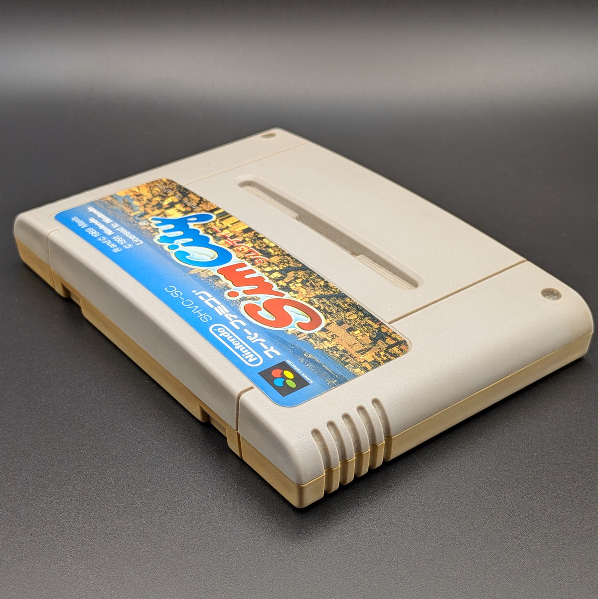 Sim City video game cartridge for Nintendo's Super Famicom (SFC) Entertainment System, shown laying on its back with the label facing upwards. Showing the left hand side and top edge. It sits on a dark grey background.