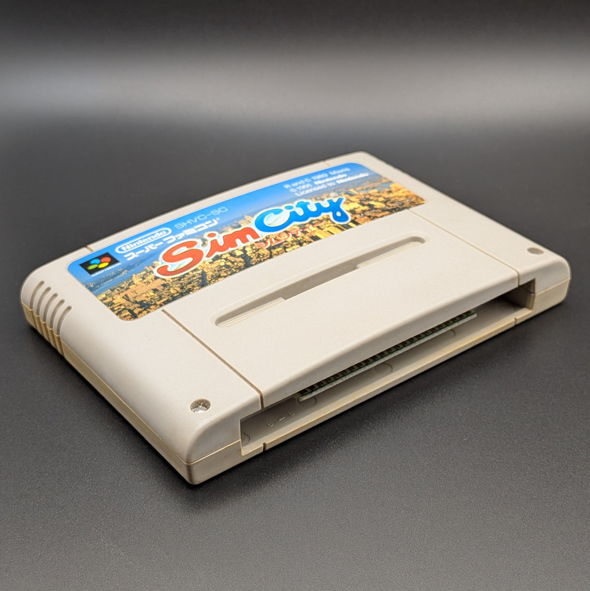 Sim City video game cartridge for Nintendo's Super Famicom (SFC) Entertainment System, shown laying on its back with the label facing upwards. Showing the left hand side and bottom slot. It sits on a dark grey background.