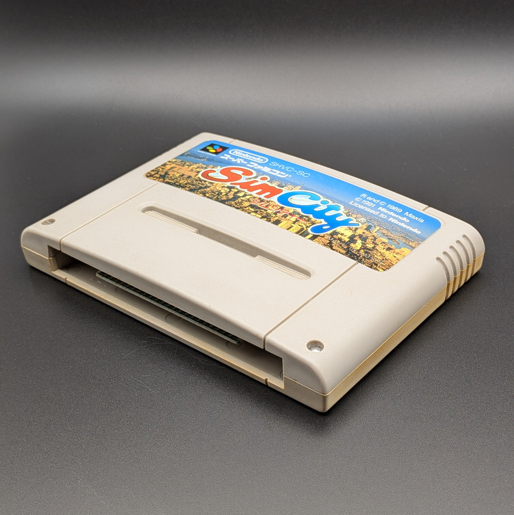 Sim City video game cartridge for Nintendo's Super Famicom (SFC) Entertainment System, shown laying on its back with the label facing upwards. Showing the right hand side and bottom slot. It sits on a dark grey background.