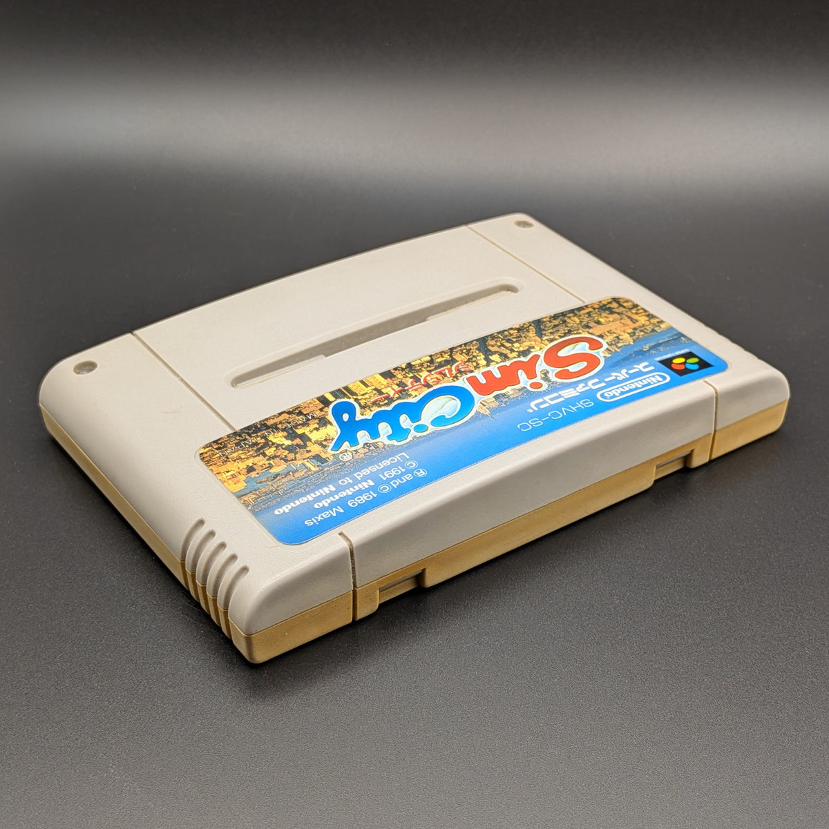 Sim City video game cartridge for Nintendo's Super Famicom (SFC) Entertainment System, shown laying on its back with the label facing upwards. Showing the right hand side and top edge. It sits on a dark grey background.