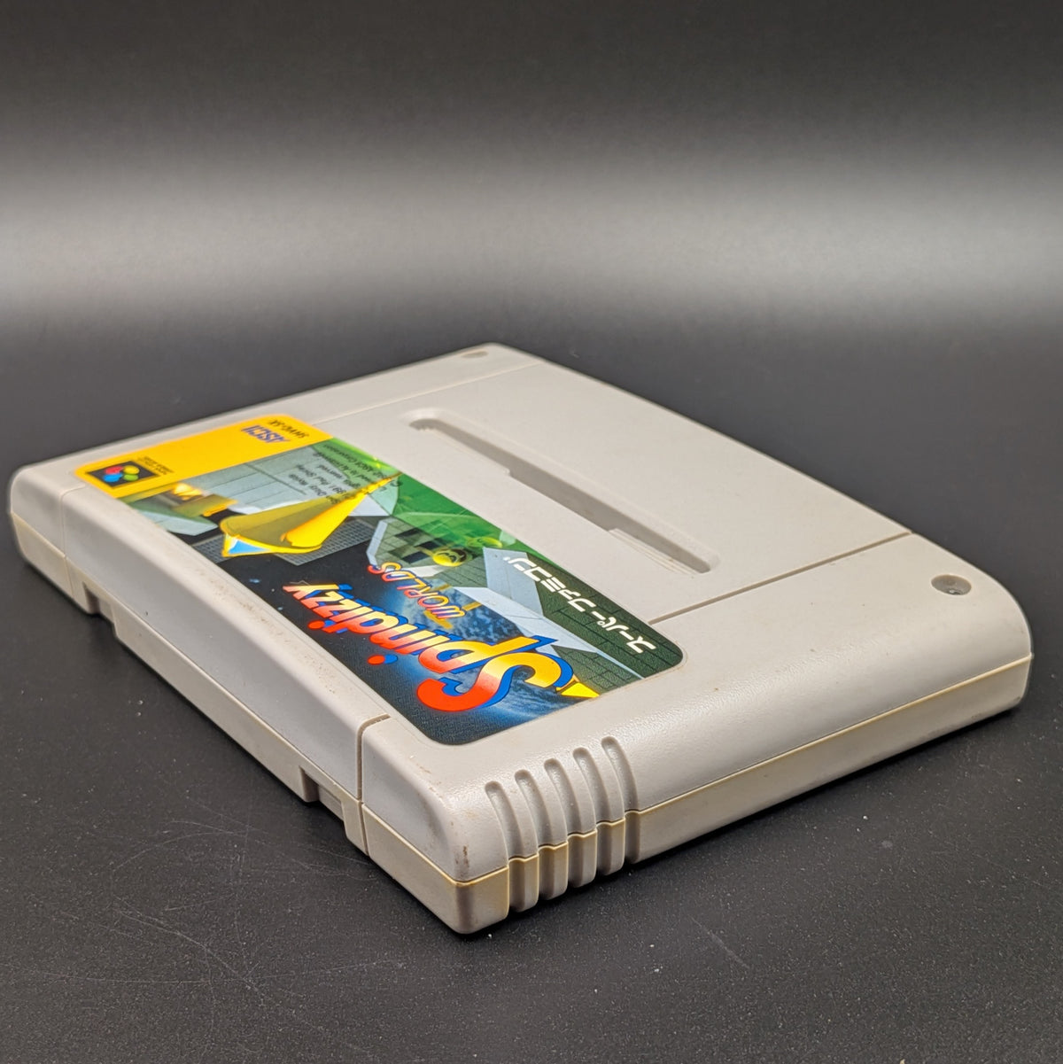 Spindizzy Worlds video game cartridge for Nintendo's Super Famicom (SFC) Entertainment System, shown laying on its back with the label facing upwards. Showing the left hand side and top edge. It sits on a dark grey background.