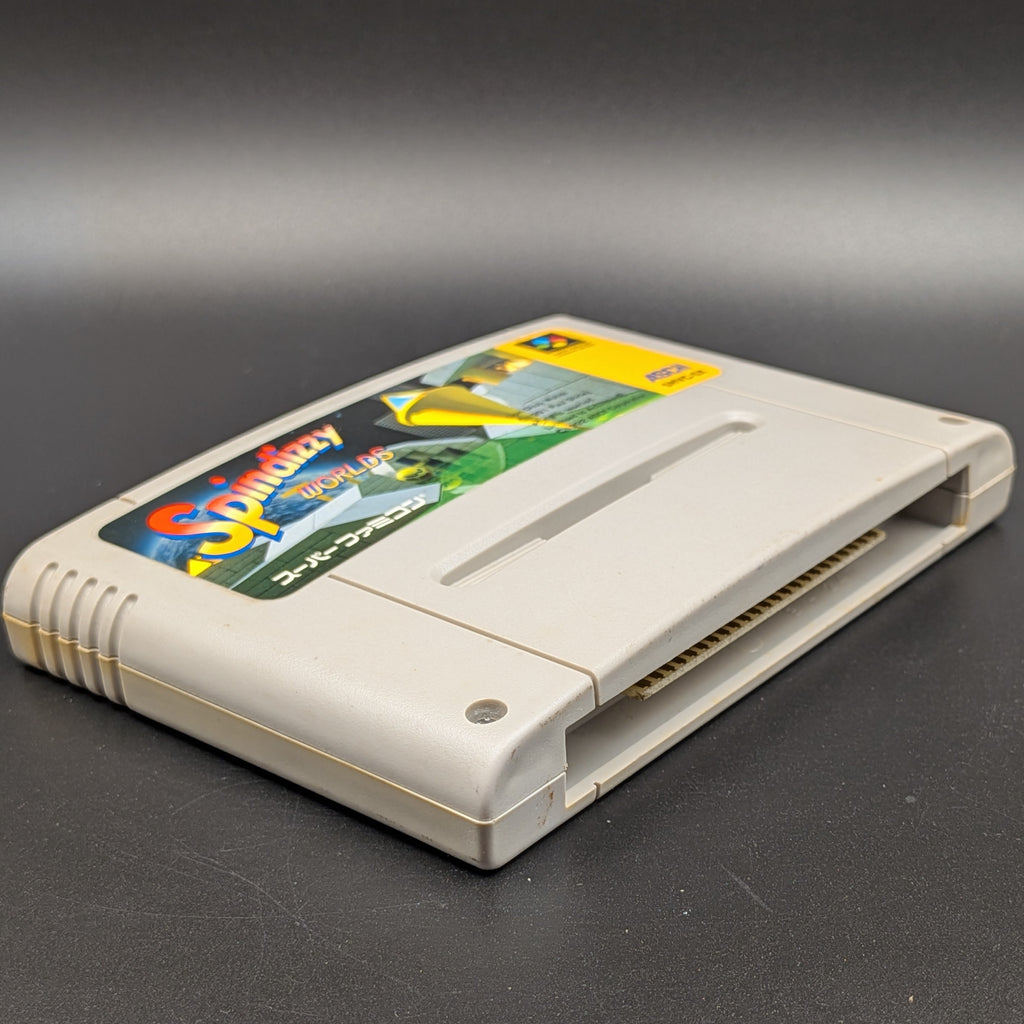 Spindizzy Worlds video game cartridge for Nintendo's Super Famicom (SFC) Entertainment System, shown laying on its back with the label facing upwards. Showing the left hand side and bottom slot. It sits on a dark grey background