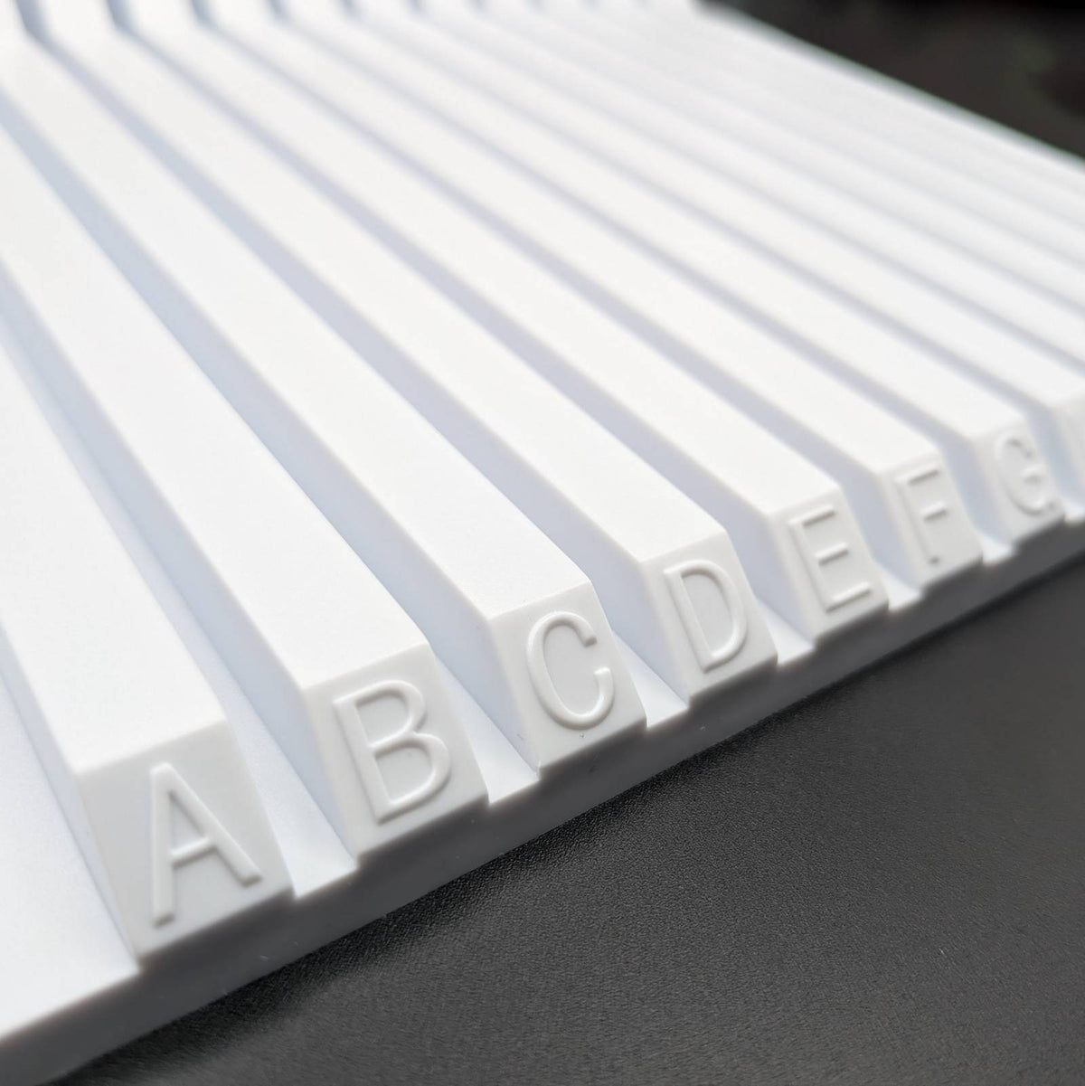 A close-up of a white alphabet letter board with raised letters A through G  visible on parallel slats, photographed on a dark gray surface.