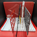 Four plastic model kit parts or accessories displayed upright on a white display stand, with a red box in the background. The parts are in red, black, silver, and yellow, arranged side by side against a red backdrop.