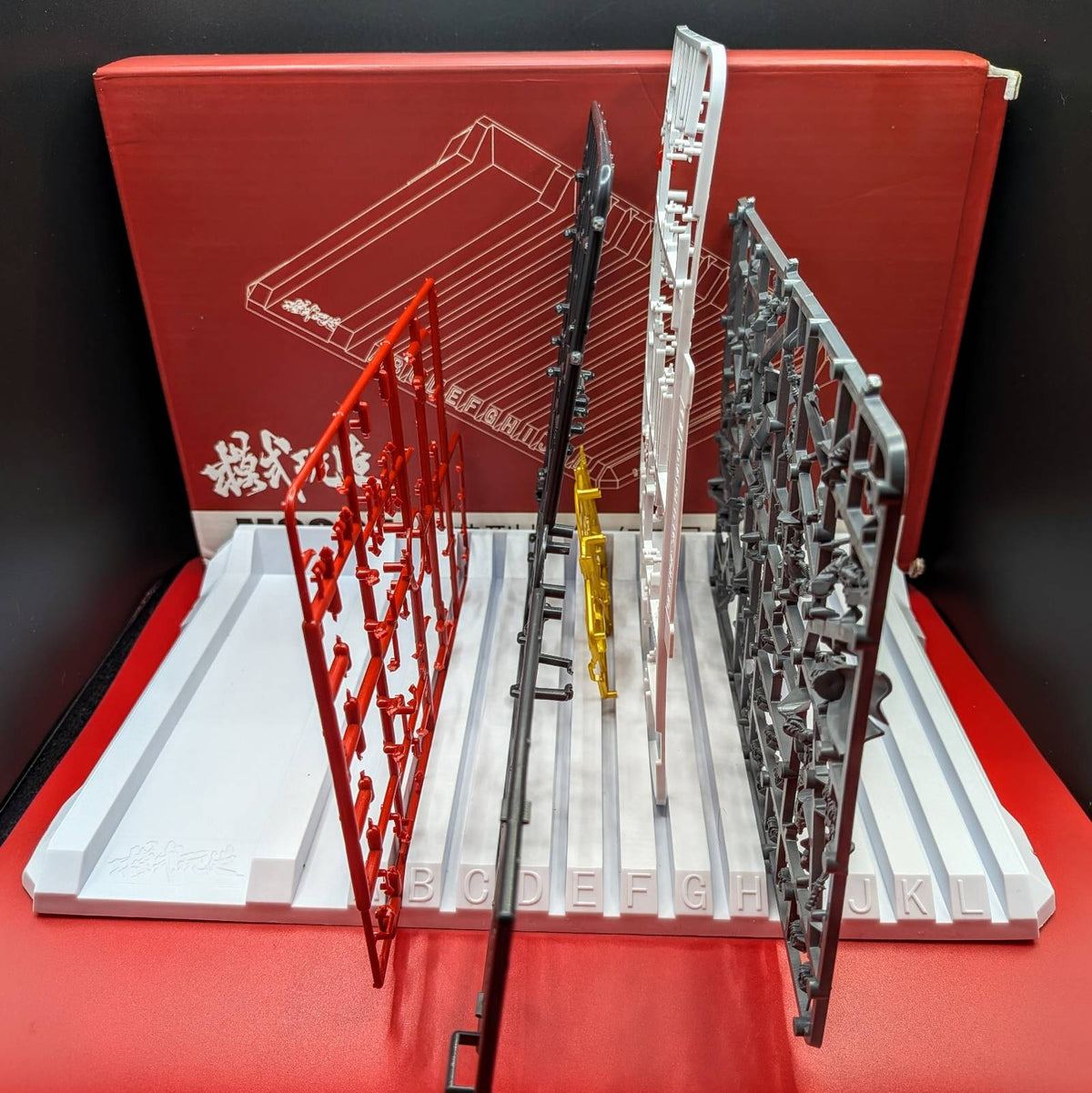 Four plastic model kit parts or accessories displayed upright on a white display stand, with a red box in the background. The parts are in red, black, silver, and yellow, arranged side by side against a red backdrop.