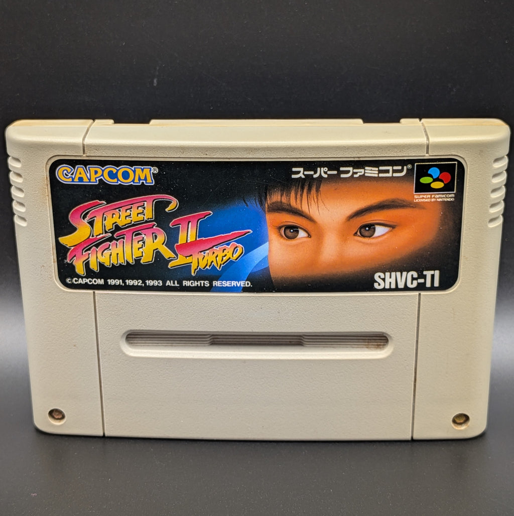A close up of Street Fighter II Turbo: Hyper Fighting video game cartridge for Nintendo's Super Famicom (SFC) Entertainment System, shown standing up and facing forwards on a dark grey background.