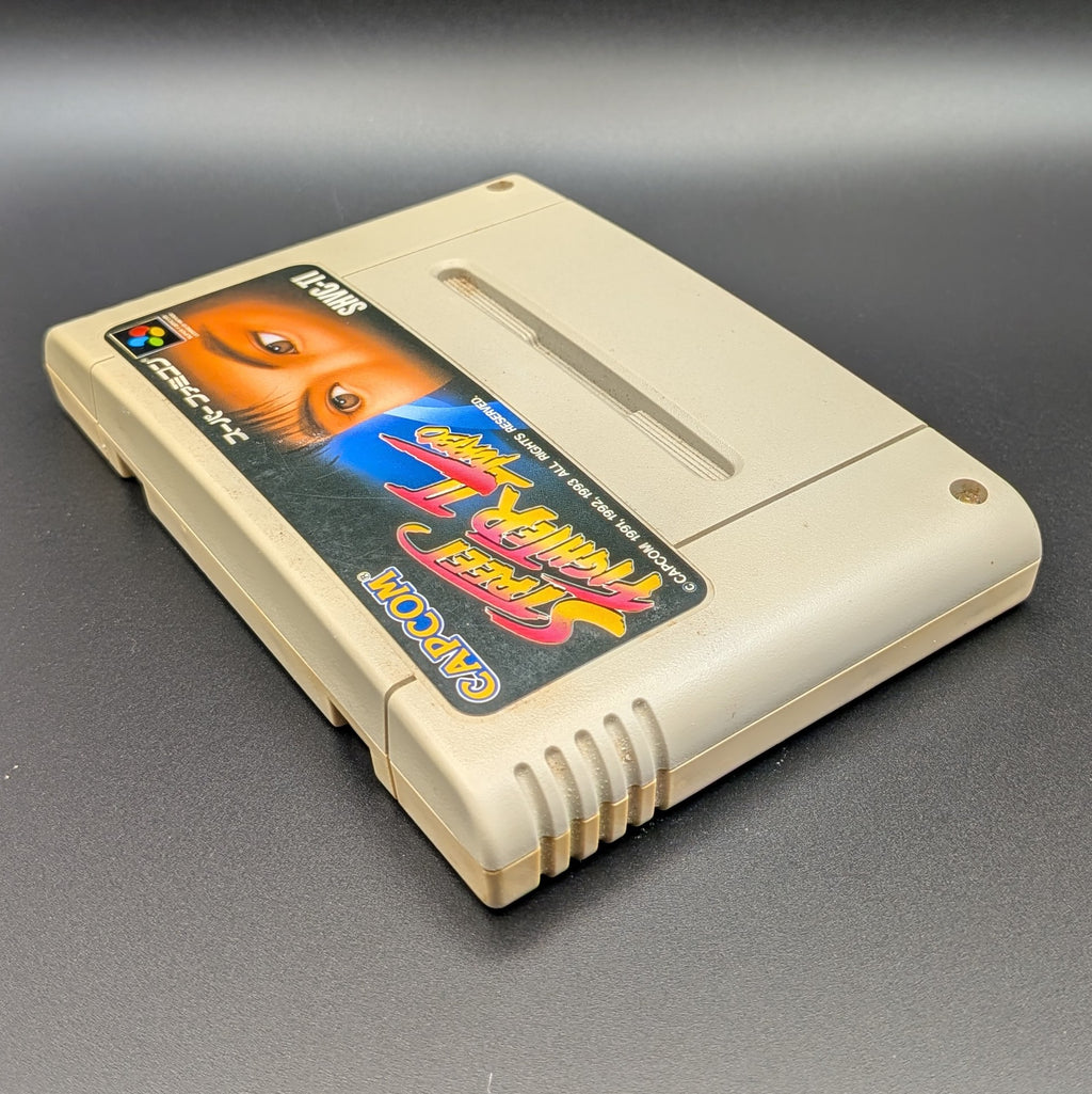Street Fighter II Turbo: Hyper Fighting video game cartridge for Nintendo's Super Famicom (SFC) Entertainment System, shown laying on its back with the label facing upwards. Showing the left hand side and top edge. It is displayed on a dark grey background.