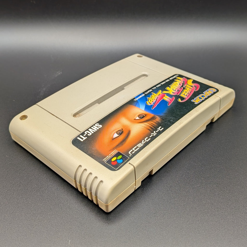 Street Fighter II Turbo: Hyper Fighting video game cartridge for Nintendo's Super Famicom (SFC) Entertainment System, shown laying on its back with the label facing upwards. Showing the right hand side and top edge. It is displayed on a dark grey background.