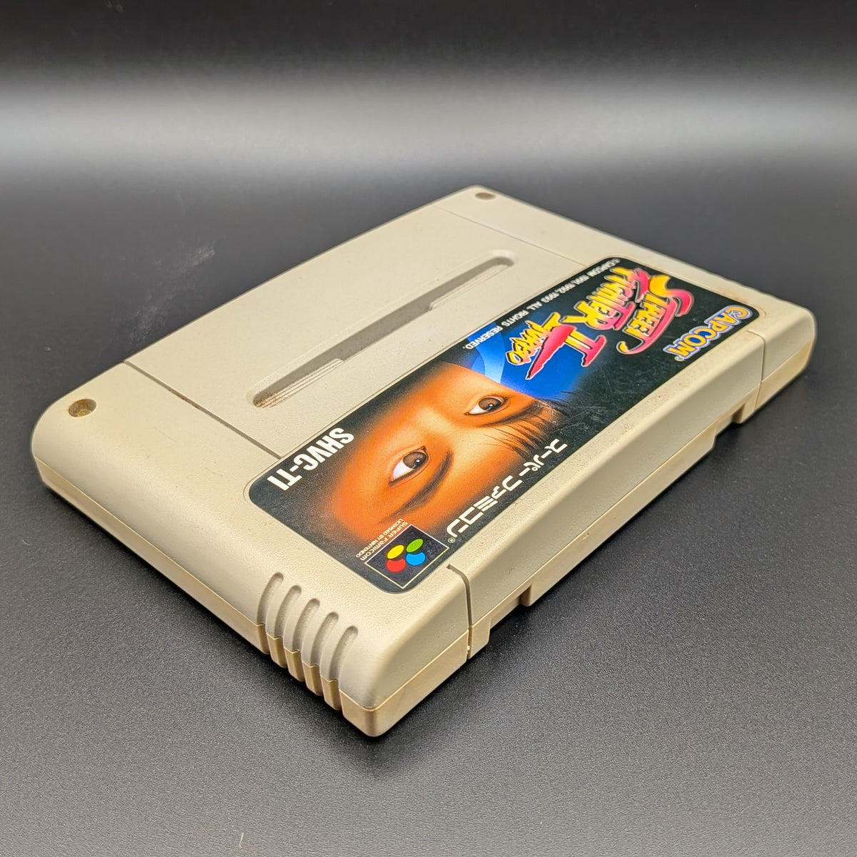 Street Fighter II Turbo: Hyper Fighting video game cartridge for Nintendo's Super Famicom (SFC) Entertainment System, shown laying on its back with the label facing upwards. Showing the right hand side and top edge. It is displayed on a dark grey background.