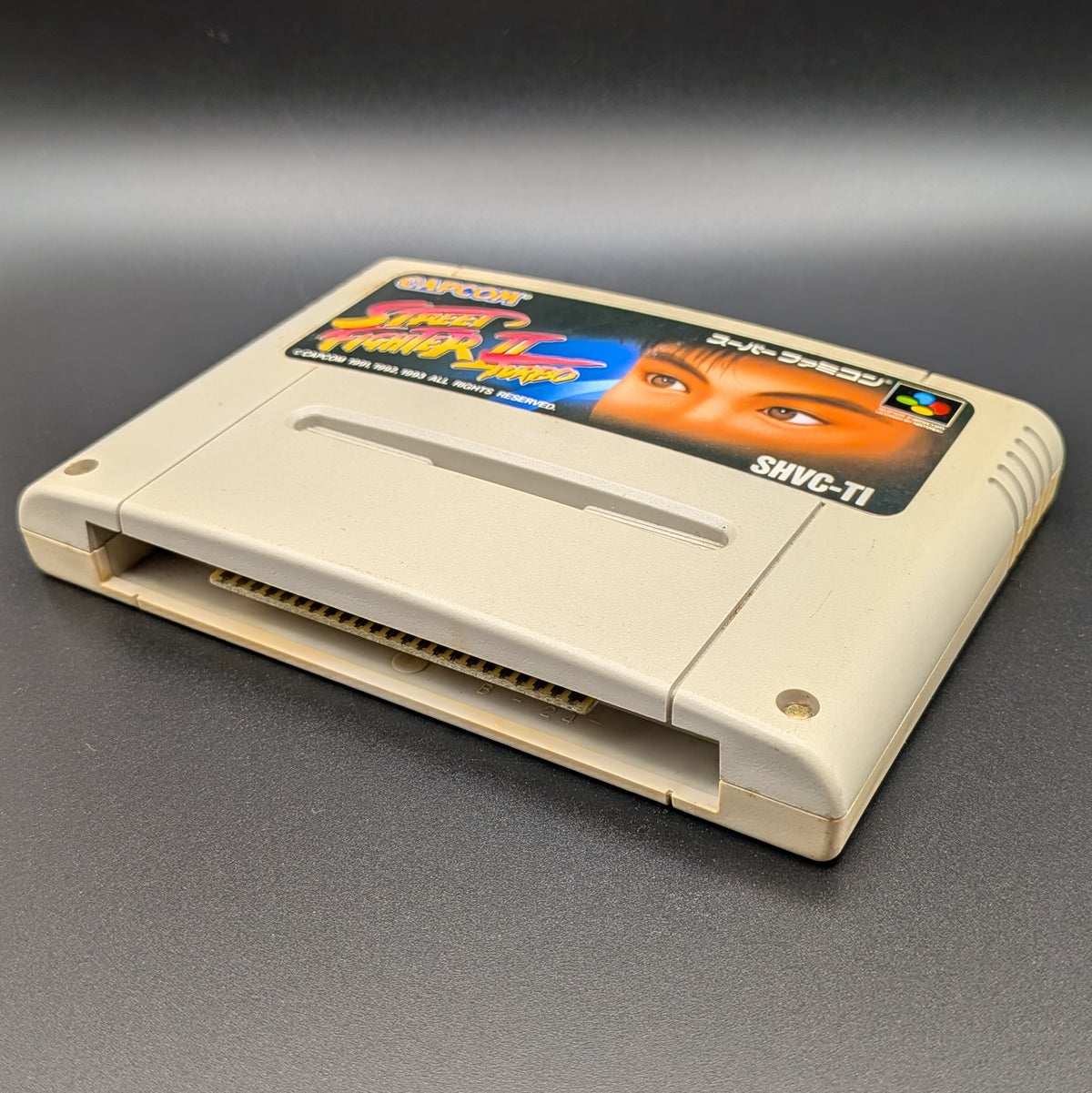 Street Fighter II Turbo: Hyper Fighting video game cartridge for Nintendo's Super Famicom (SFC) Entertainment System, shown laying on its back with the label facing upwards. Showing the right hand side and bottom slot. It is displayed on a dark grey background.