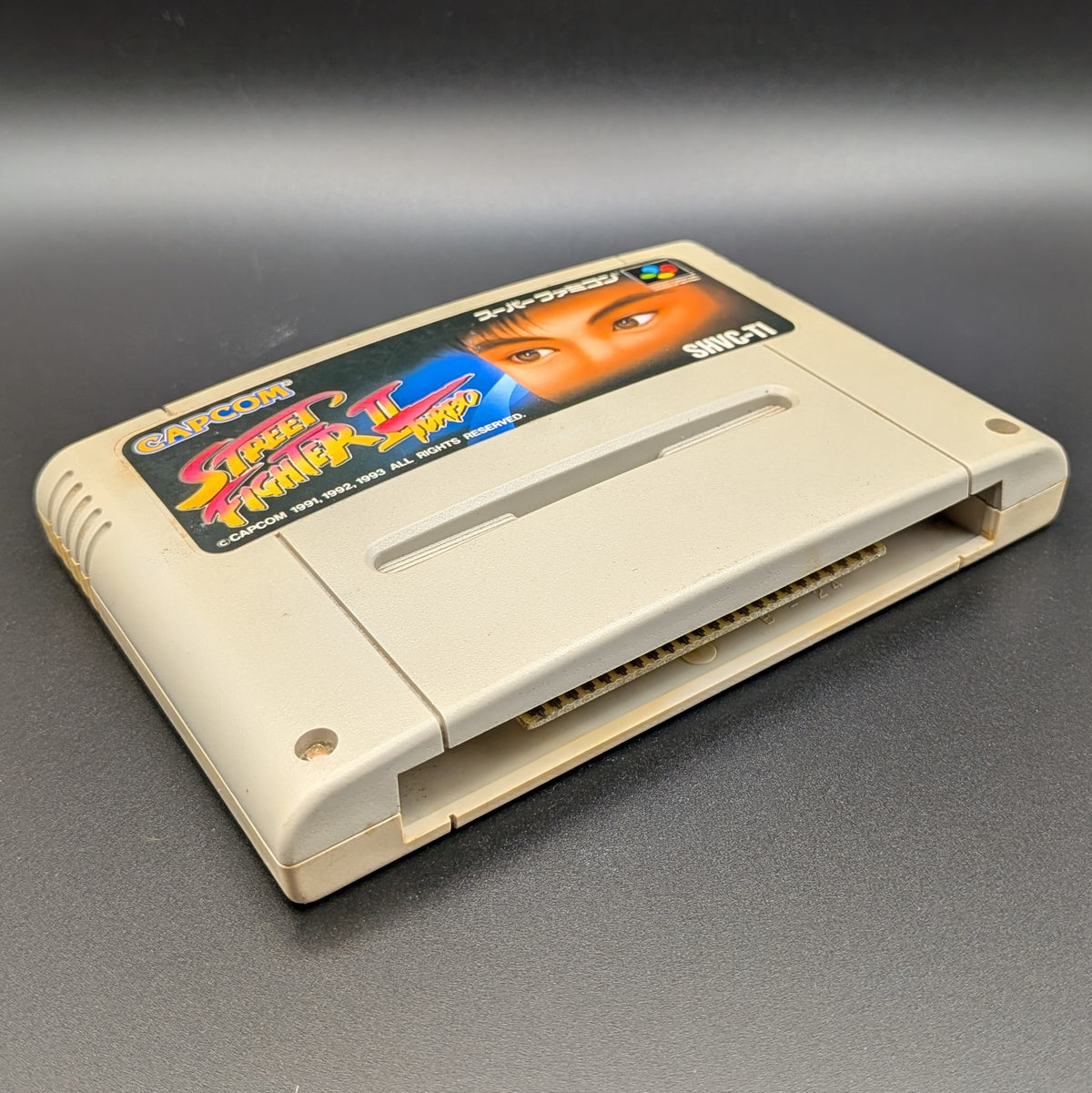 Street Fighter II Turbo: Hyper Fighting video game cartridge for Nintendo's Super Famicom (SFC) Entertainment System, shown laying on its back with the label facing upwards. Showing the left hand side and bottom slot. It is displayed on a dark grey background.