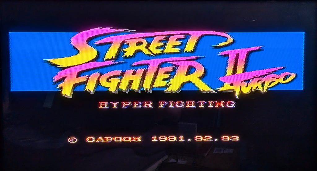 Street Fighter II Turbo : Hyper Fighting title game screen. A retro neon-style text, on a dark background, with the title in pink and yellow lettering and Capcom copyright text below. Released by Nintendo for the Super Famicom (SFC).