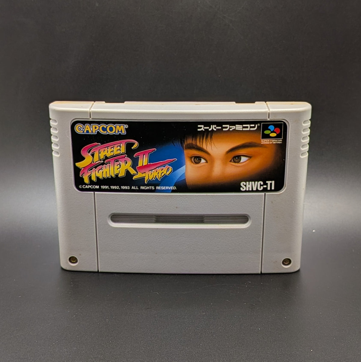 Street Fighter 2 Turbo: Hyper Fighting video game cartridge for the Super Famicom Entertainment System, shown standing up and facing forward on a dark grey background.
