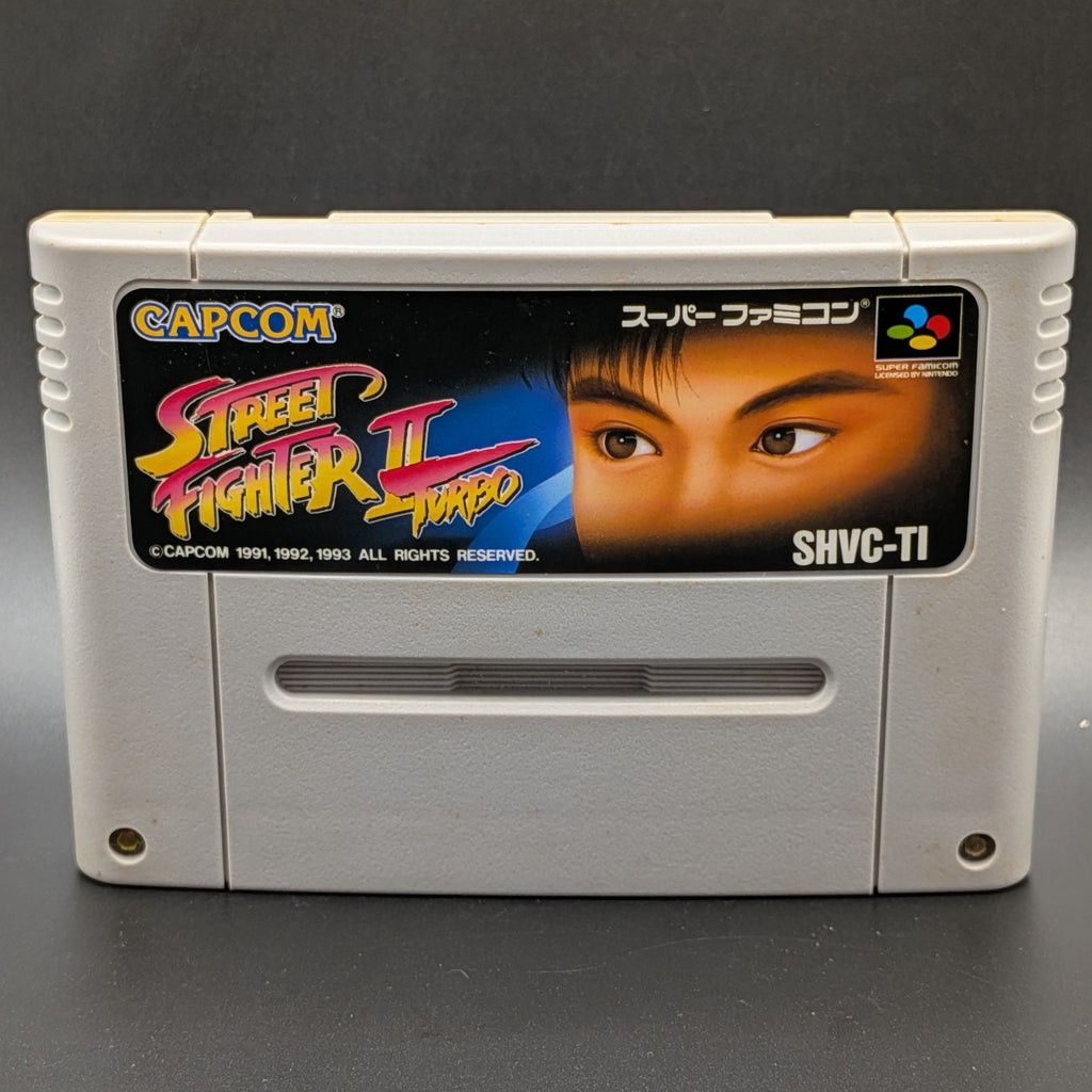 A close up of Street Fighter 2 Turbo: Hyper Fighting video game cartridge for the Super Famicom Entertainment System, shown standing up and facing forward on a dark grey background.
