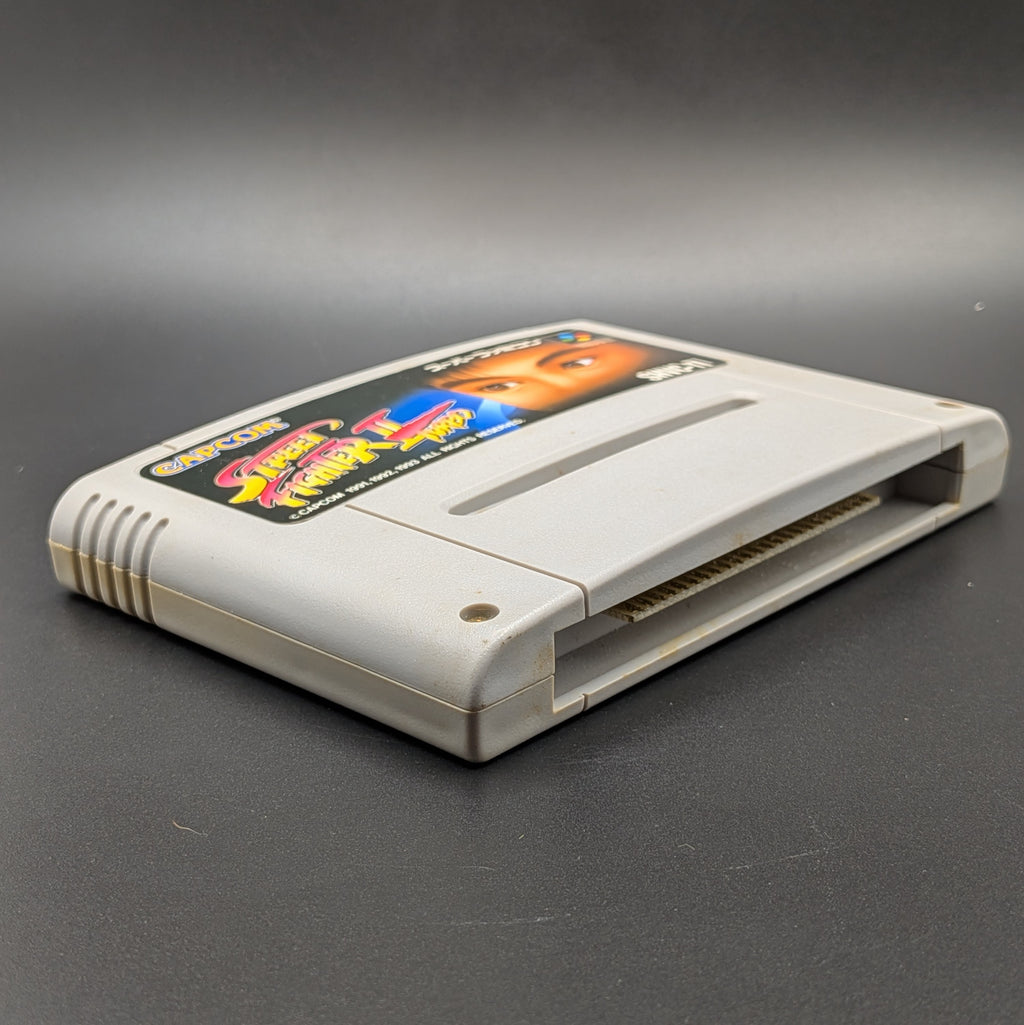 Street Fighter 2 Turbo: Hyper Fighting video game cartridge for the Super Famicom Entertainment System, shown laying on its back with the label facing up, at a 3/4 angle. Pictured on a dark grey background.