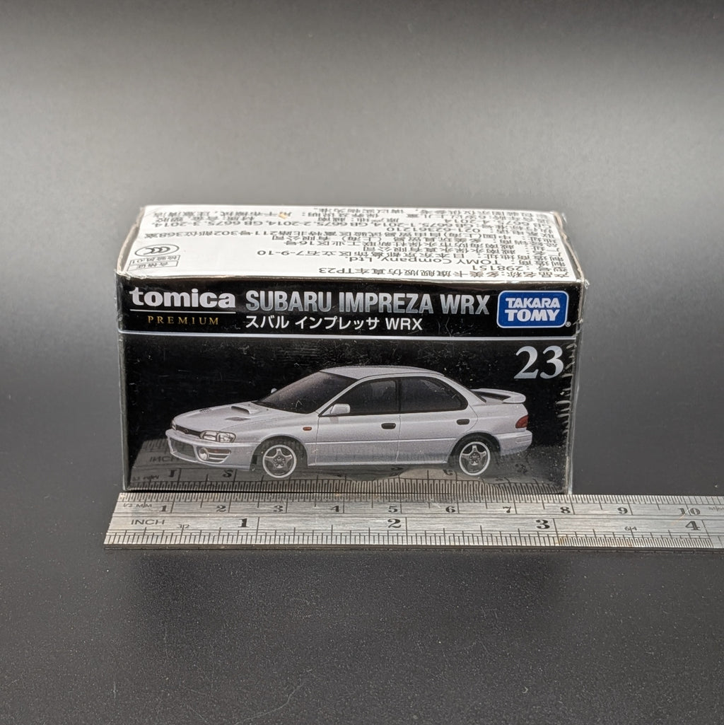A Takara Tomy model car sealed in its packaging. This Tomica premium model shows a Subaru Impreza WRX on the front of the packaging which is sat on a dark grey background, with a ruler running across the bottom for scale.