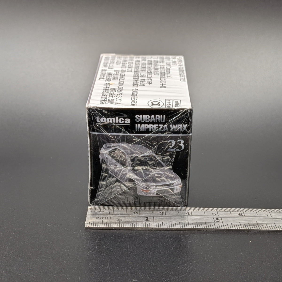 A Takara Tomy model car sealed in its packaging. Shown from the side, with a ruler for scale, sat on a dark grey background.
