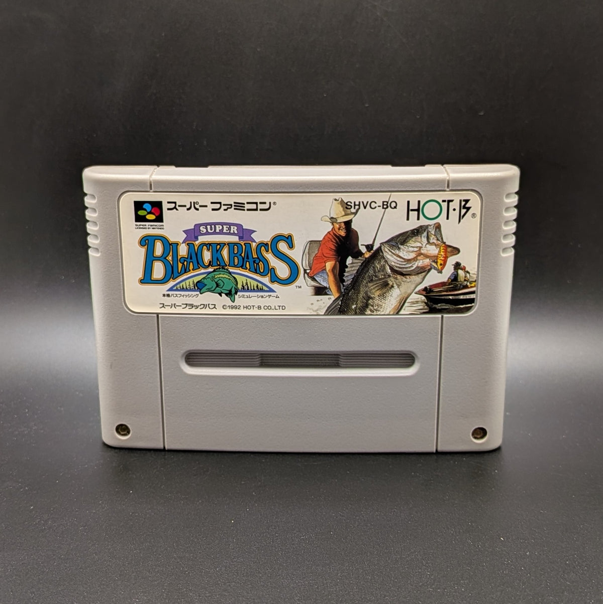 Super Black Bass video game cartridge for Nintendo's Super Famicom (SFC) Entertainment System, shown standing up and facing forward on a dark grey background.
