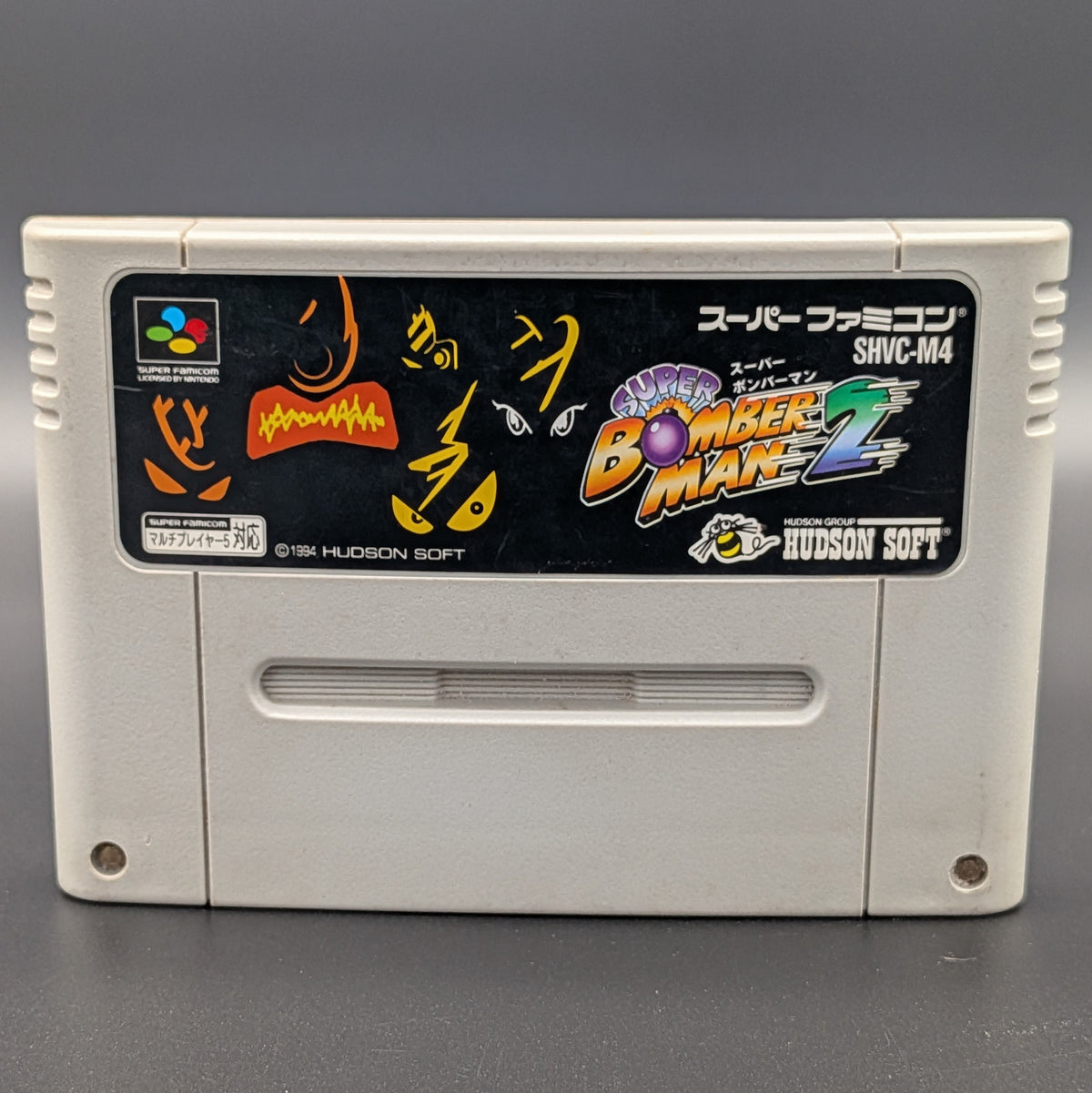 A close up of Super Bomberman 2 video game cartridge for Nintendo's Super Famicom (SFC) Entertainment System, shown standing up and facing forwards on a dark grey background.