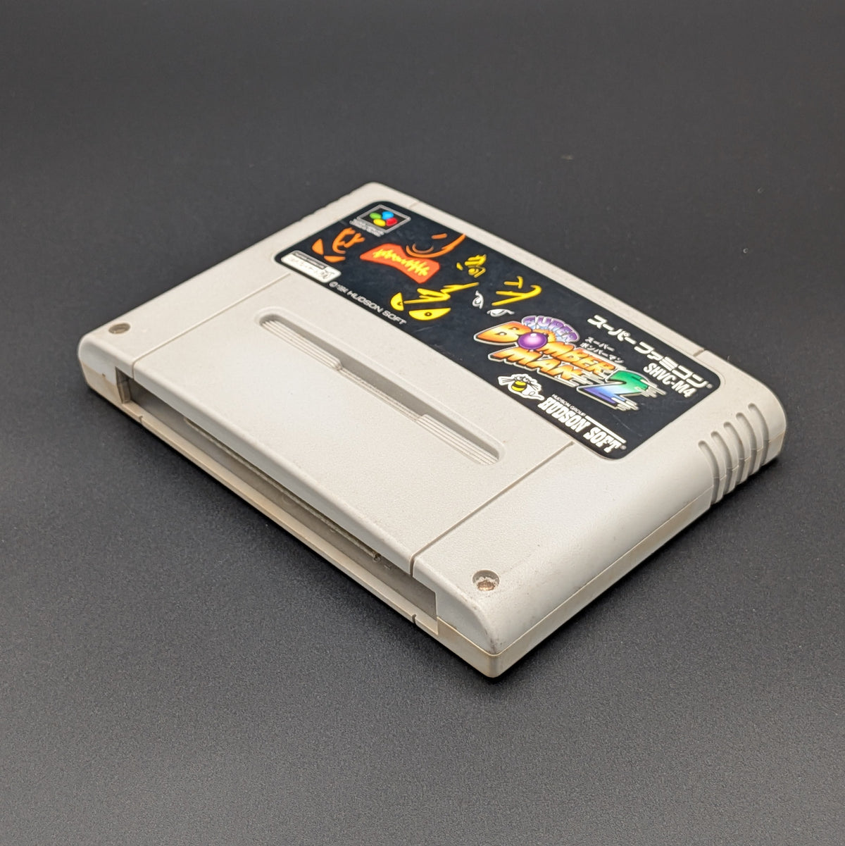 Super Bomberman 2 video game cartridge for Nintendo's Super Famicom (SFC) Entertainment System, shown laying on its back with the label facing upwards. Showing the right hand side and bottom slot. It is displayed on a dark grey background.
