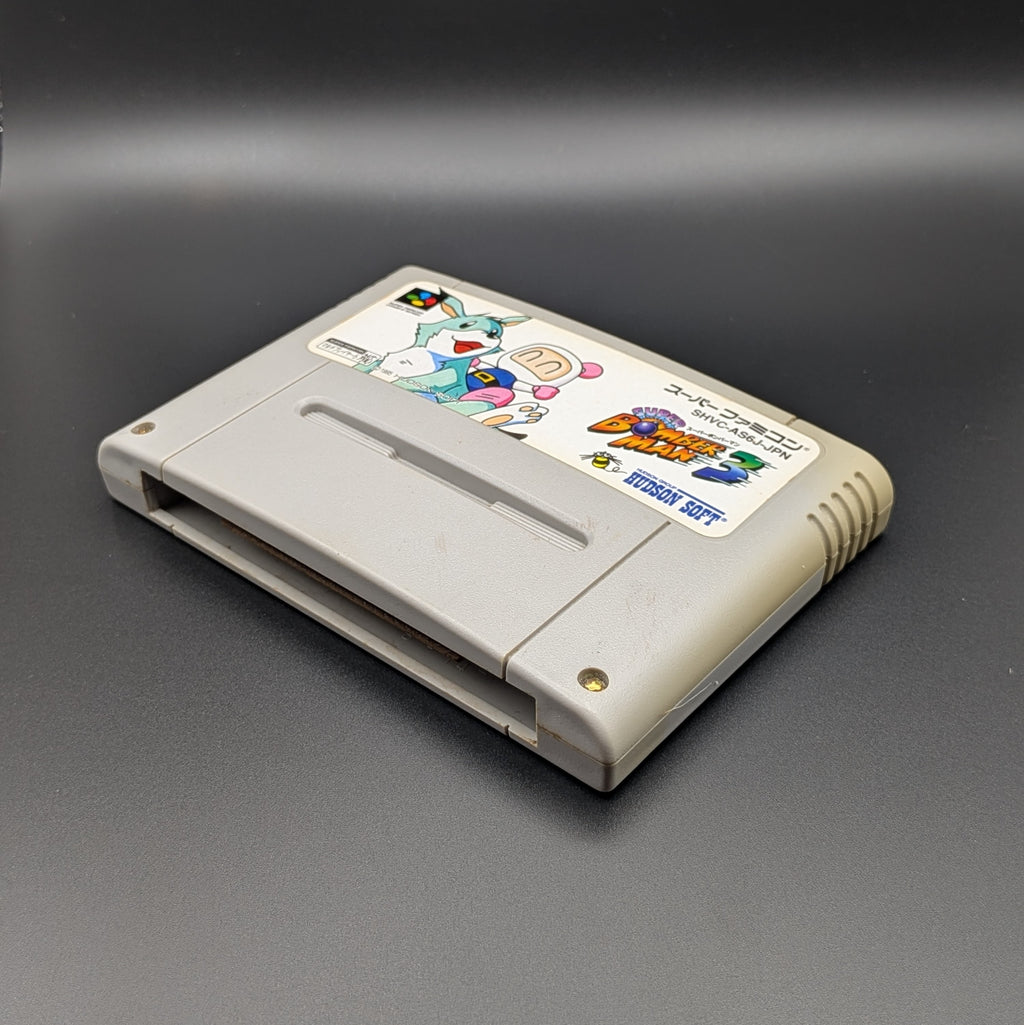 Super Bomberman 3 video game cartridge for Nintendo's Super Famicom (SFC) Entertainment System, shown laying on its back with the label facing upwards. Showing the right hand side and bottom slot. It is displayed on a dark grey background.