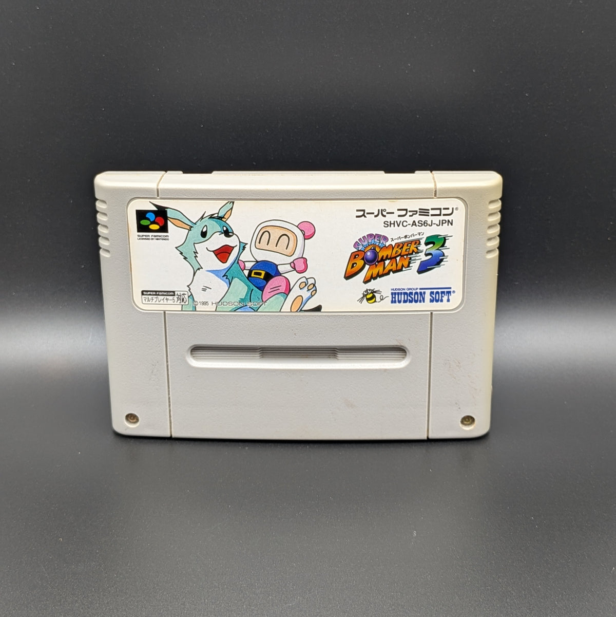 Super Bomberman 3 video game cartridge for Nintendo's Super Famicom (SFC) Entertainment System, shown standing up and facing forwards on a dark grey background.