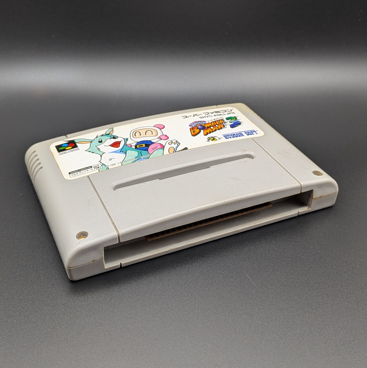 Super Bomberman 3 video game cartridge for Nintendo's Super Famicom (SFC) Entertainment System, shown laying on its back with the label facing upwards. Showing the left hand side and bottom slot. It is displayed on a dark grey background.