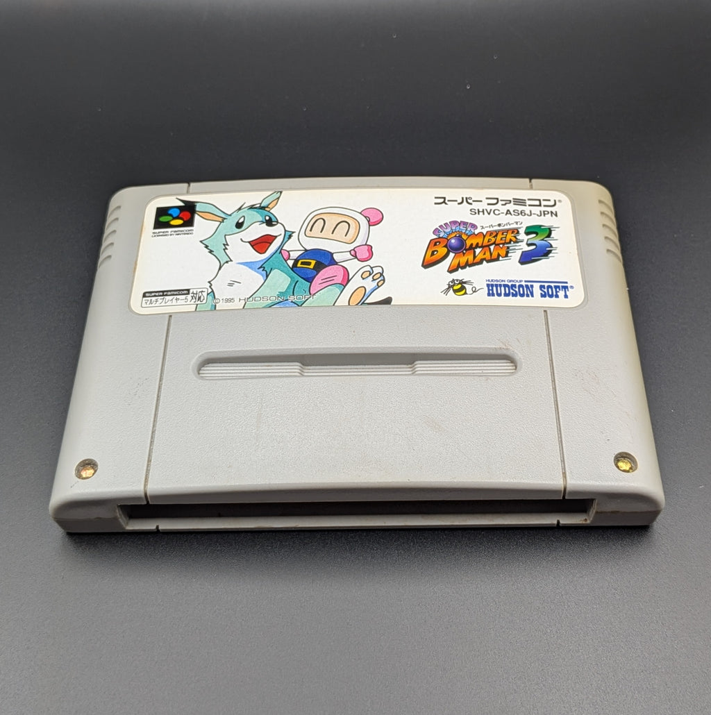 A close up of Super Bomberman 3 video game cartridge for Nintendo's Super Famicom (SFC) Entertainment System, shown laying down and facing forwards on a dark grey background.