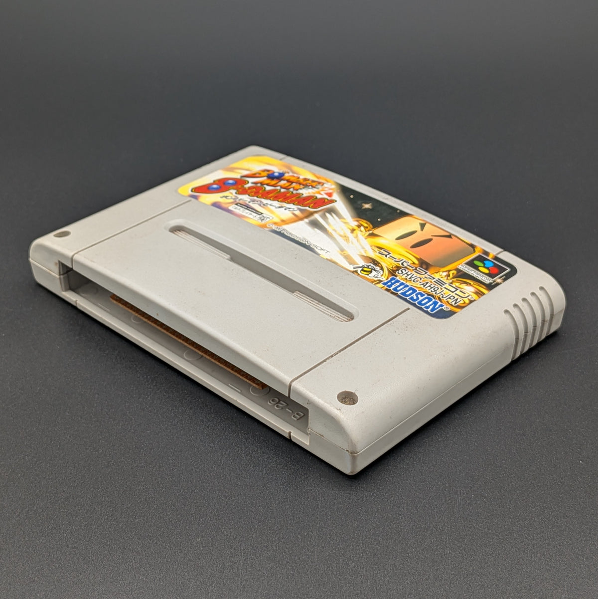 Super Bomberman B-Daman video game cartridge for Nintendo's Super Famicom (SFC) Entertainment System, shown laying on its back with the label facing upwards. It sits on a dark grey background.

