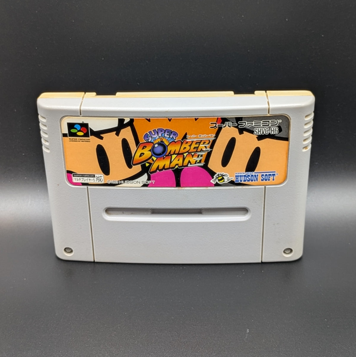 Super Bomberman video game cartridge for Nintendo's Super Famicom (SFC) Entertainment System, shown standing up and facing forwards on a dark grey background.