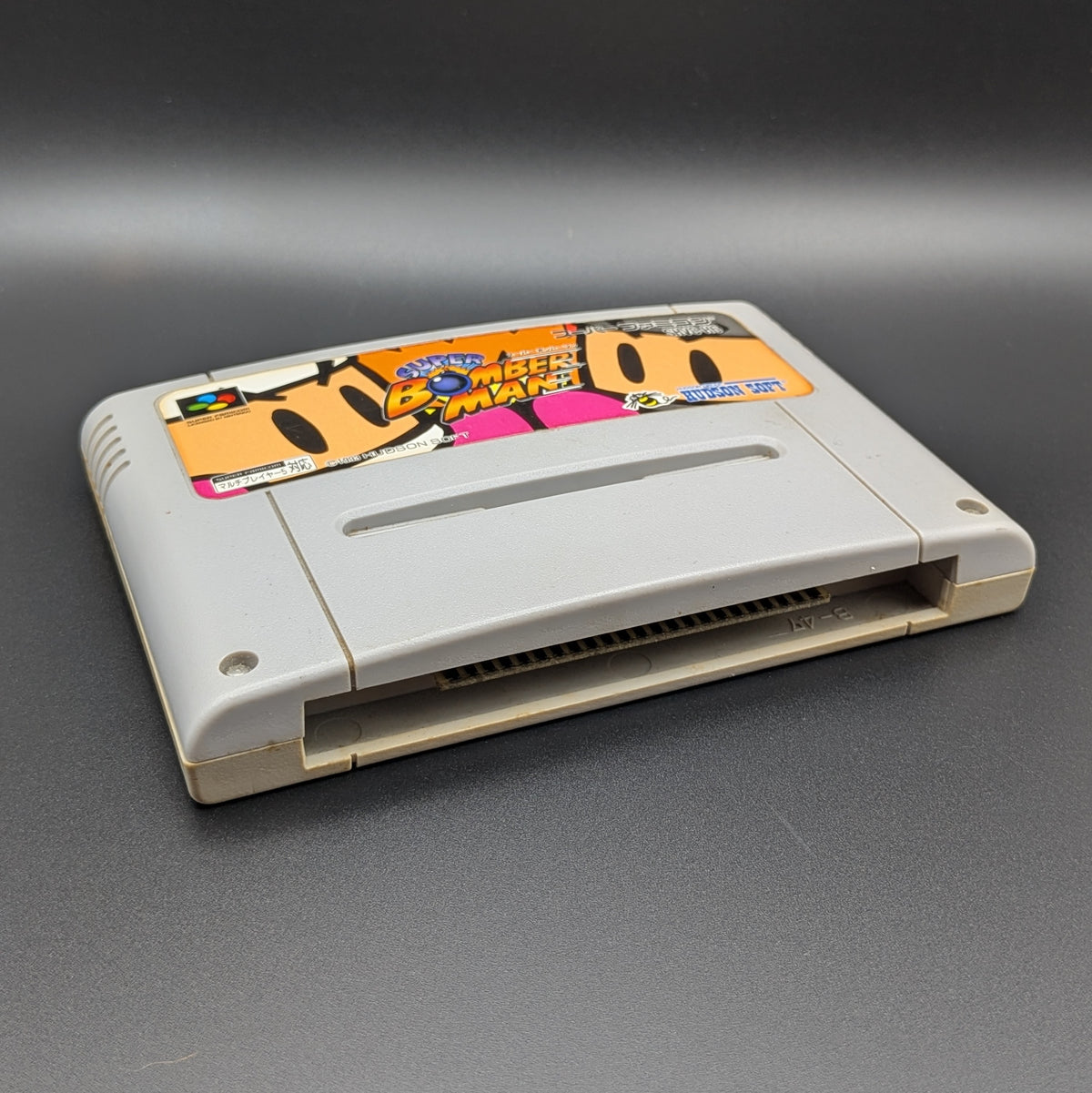 Super Bomberman video game cartridge for Nintendo's Super Famicom (SFC) Entertainment System, shown laying on its back with the label facing upwards. Showing the left hand side and bottom slot. It is displayed on a dark grey background.