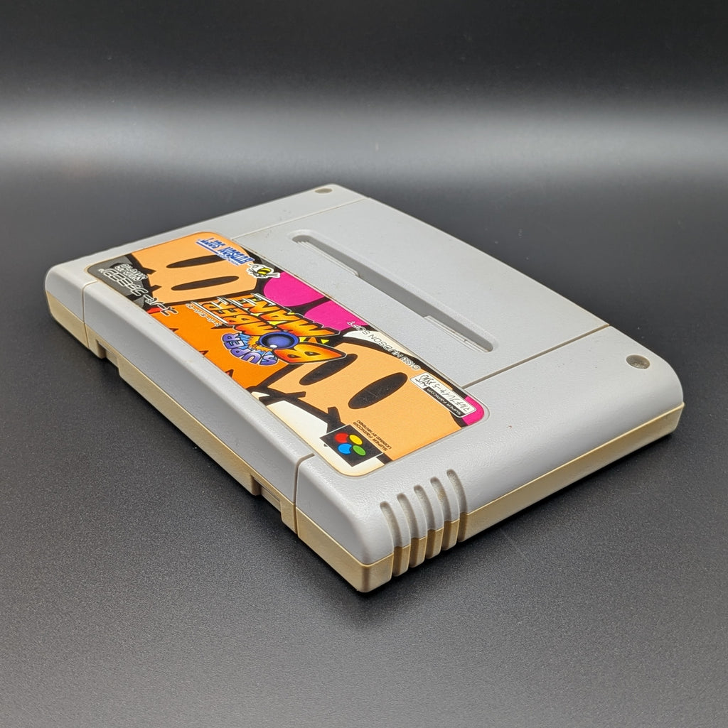 Super Bomberman video game cartridge for Nintendo's Super Famicom (SFC) Entertainment System, shown laying on its back with the label facing upwards. Showing the right hand side and top edge. It is displayed on a dark grey background.