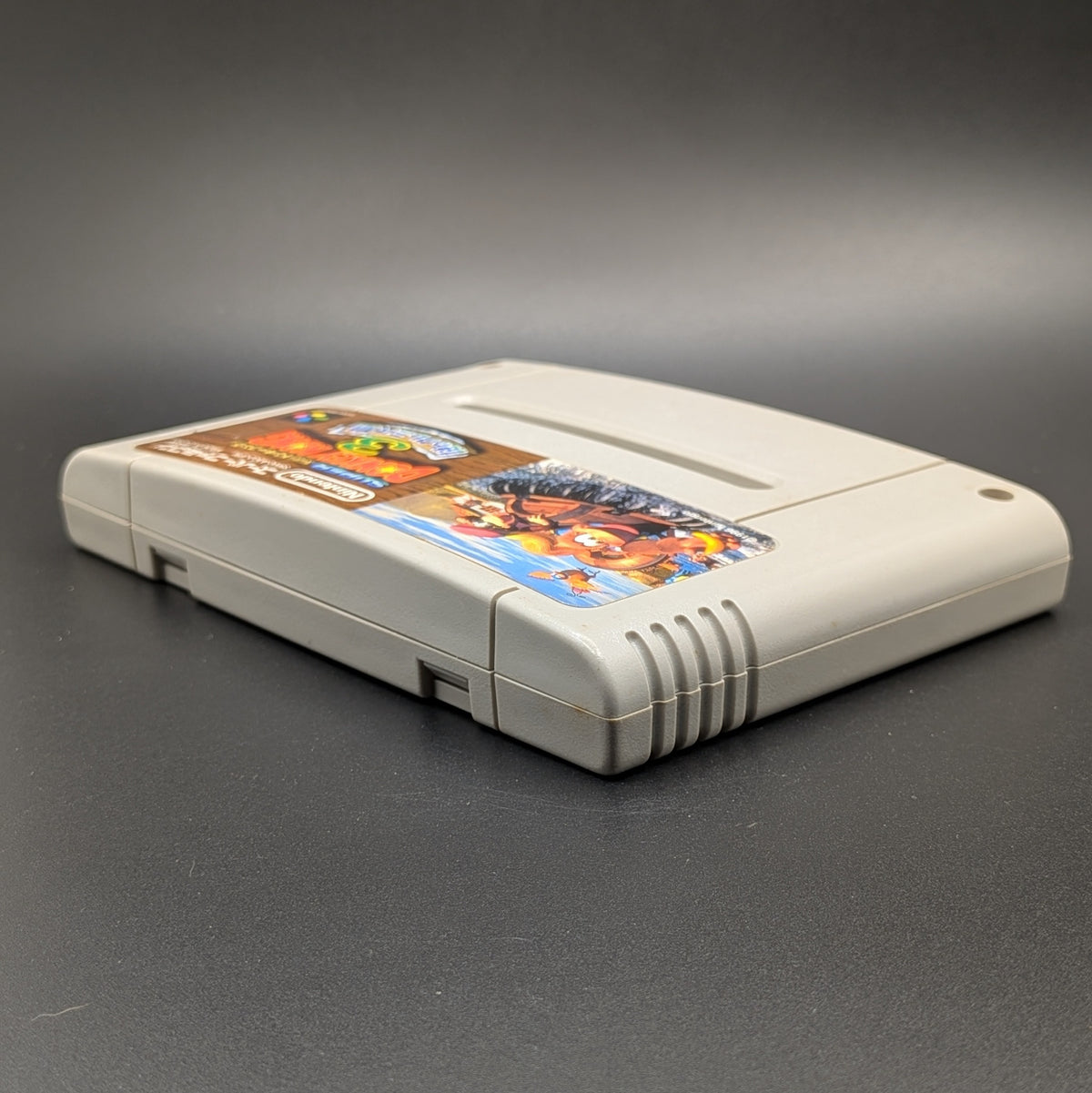 Super Donkey Kong 3 video game cartridge for Nintendo's Super Famicom (SFC) Entertainment System, shown laying on its back with the label facing up on a dark grey background.
