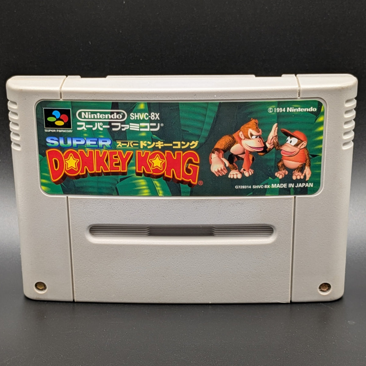 A close up of Super Donkey Kong video game cartridge for Nintendo's Super Famicom (SFC) Entertainment System, shown standing up and facing forward on a dark grey background.
