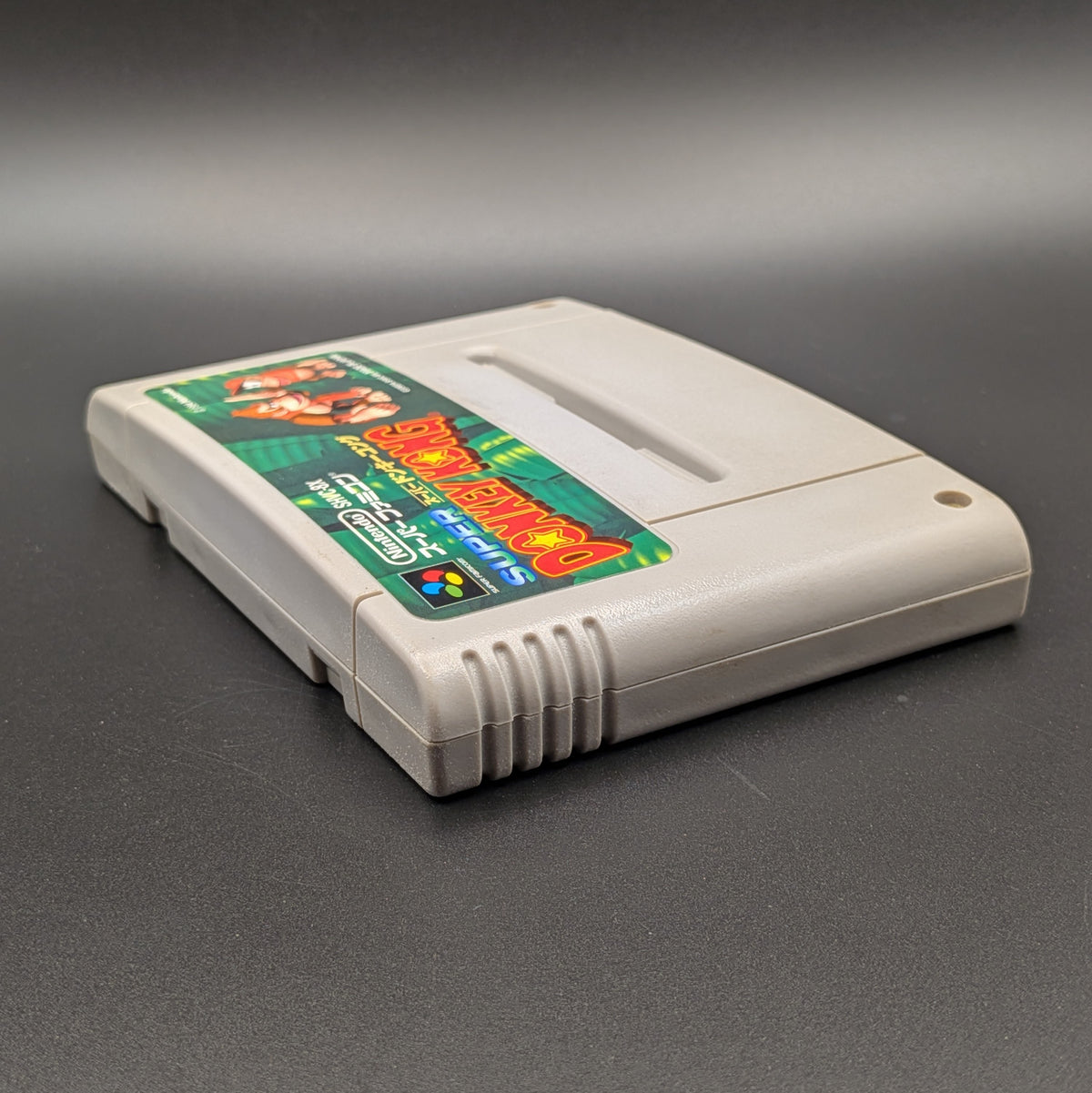 Super Donkey Kong video game cartridge for Nintendo's Super Famicom (SFC) Entertainment System, shown laying on its back with the label facing upwards. Showing the left hand side and top edge. It sits on a dark grey background.