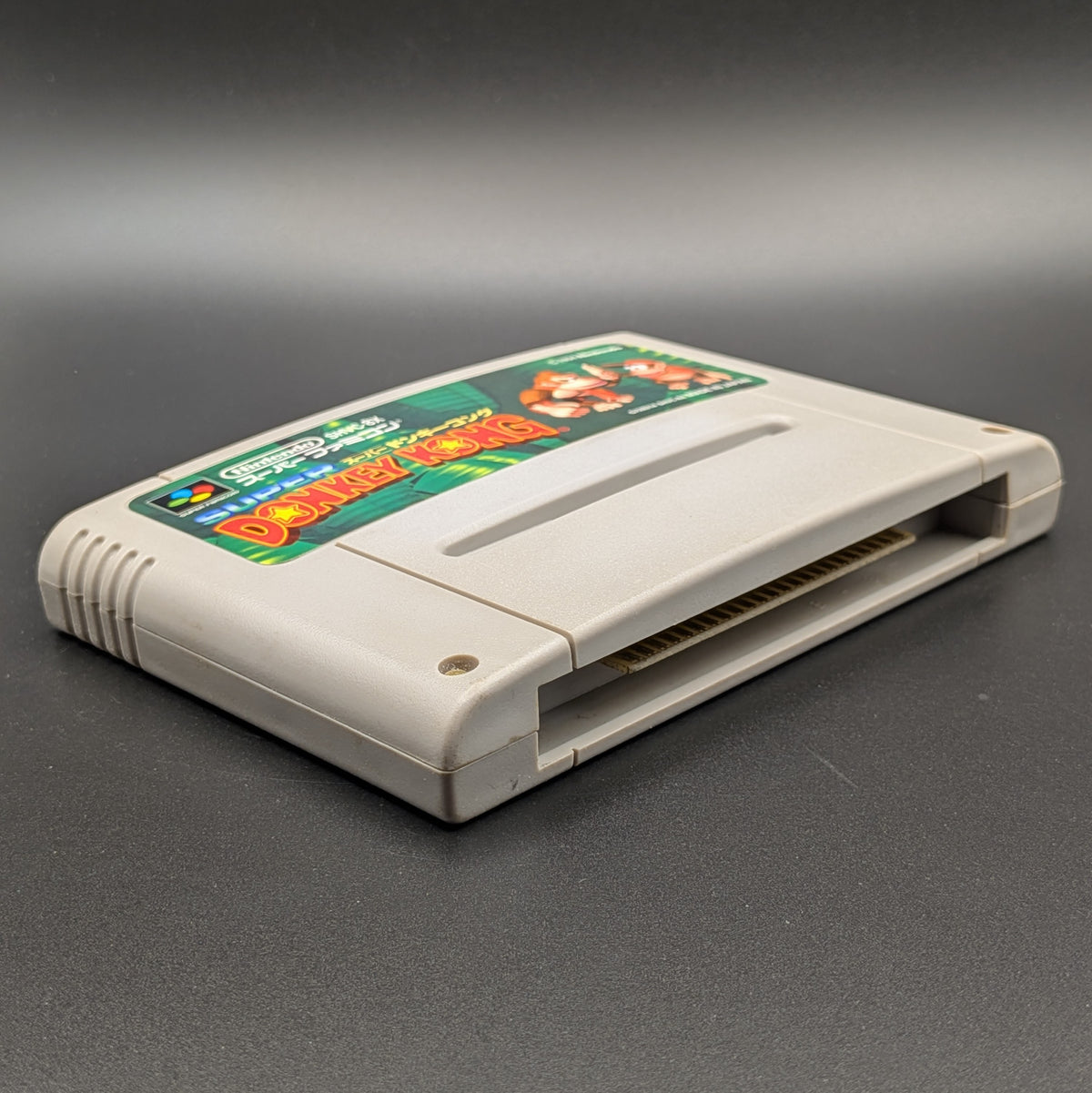 Super Donkey Kong video game cartridge for Nintendo's Super Famicom (SFC) Entertainment System, shown laying on its back with the label facing upwards. Showing the left hand side and bottom slot. It sits on a dark grey background.