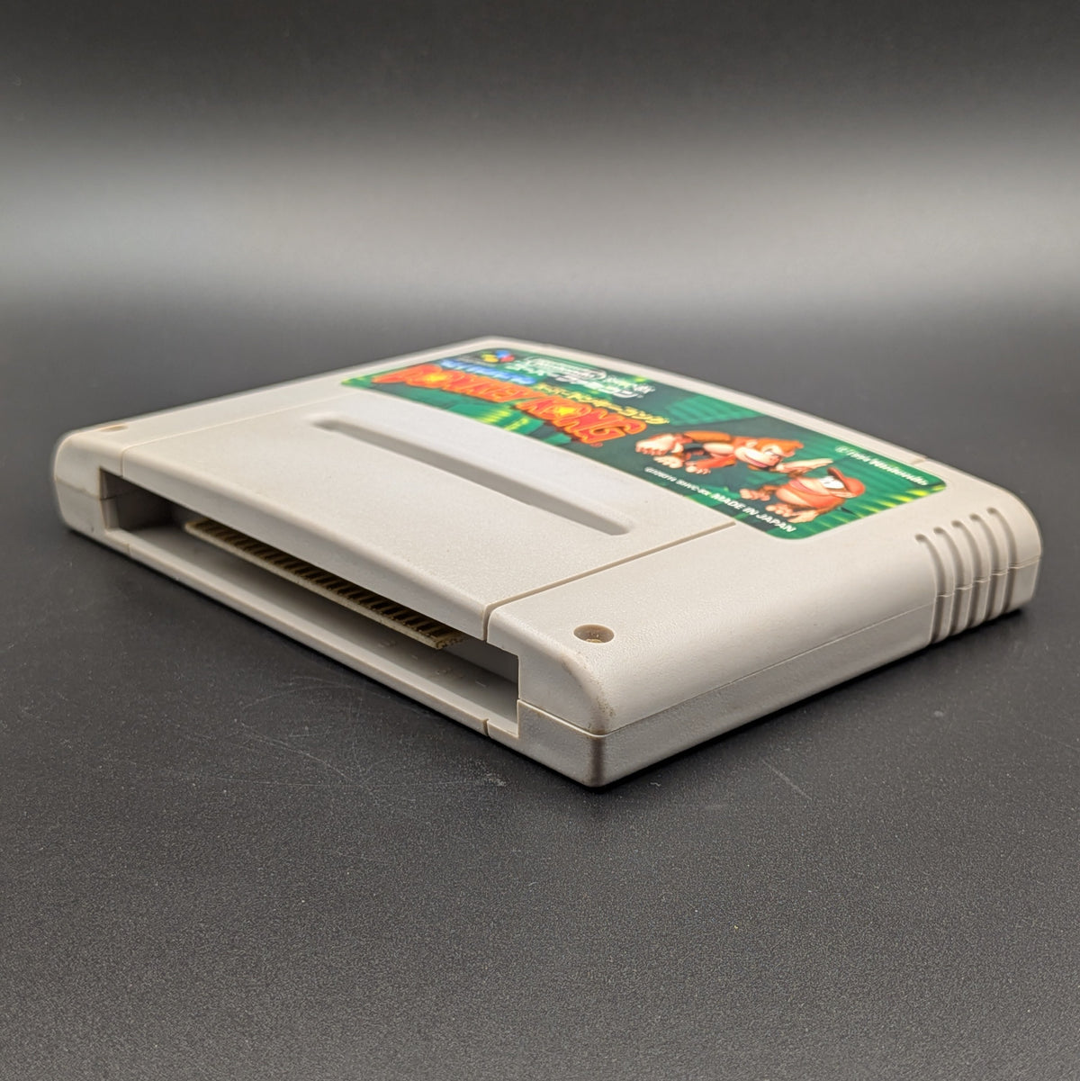 Super Donkey Kong video game cartridge for Nintendo's Super Famicom (SFC) Entertainment System, shown laying on its back with the label facing upwards. Showing the right hand side and bottom slot. It sits on a dark grey background.