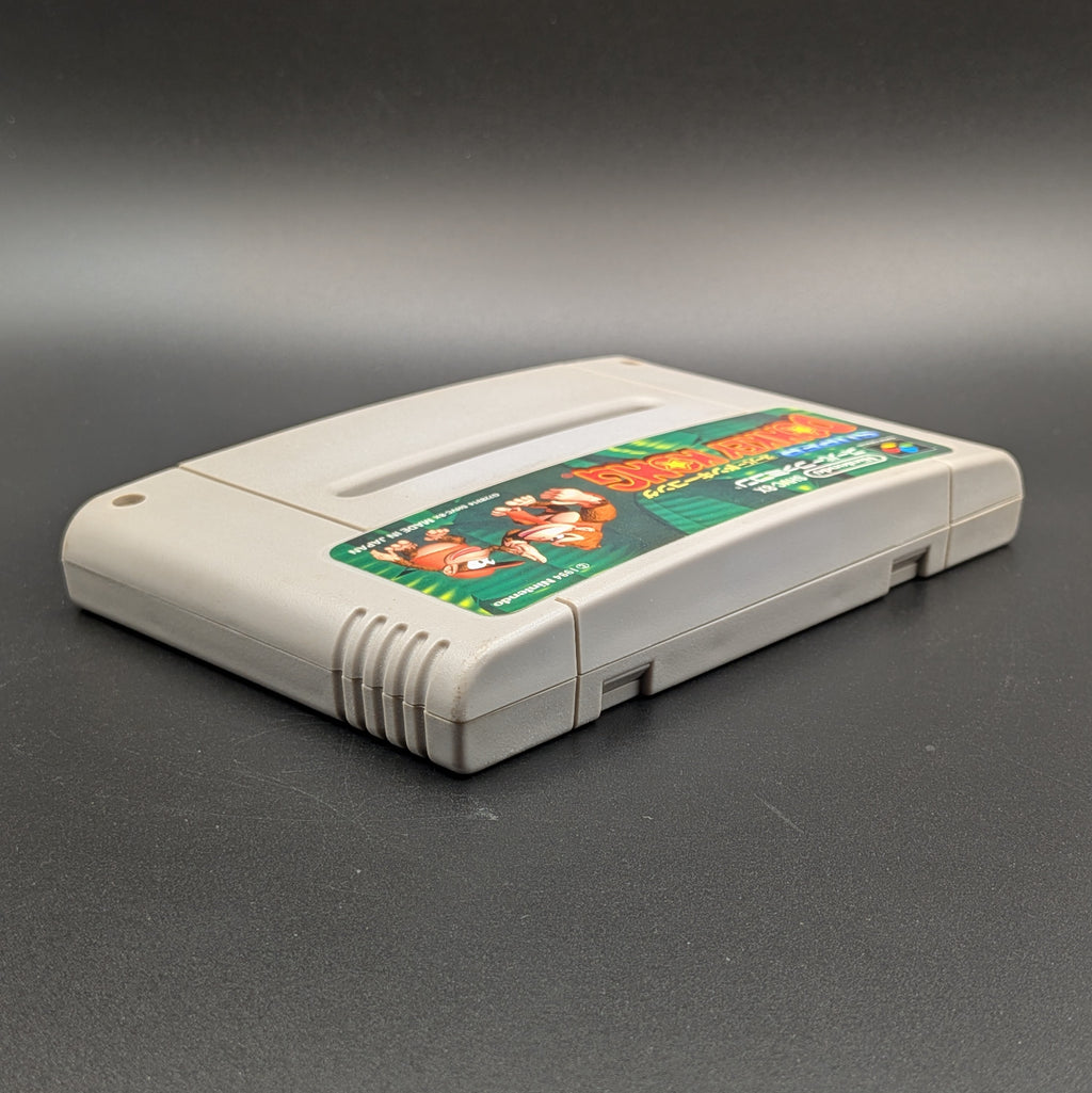 Super Donkey Kong video game cartridge for Nintendo's Super Famicom (SFC) Entertainment System, shown laying on its back with the label facing upwards. Showing the right hand side and top edge. It sits on a dark grey background.