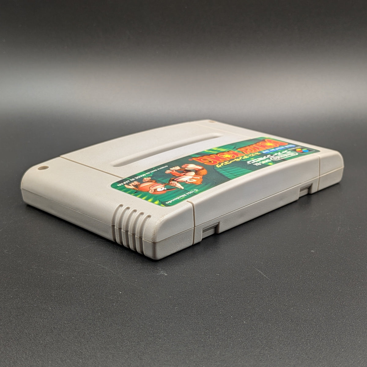 Super Donkey Kong video game cartridge for Nintendo's Super Famicom (SFC) Entertainment System, shown laying on its back with the label facing upwards. Showing the right hand side and top edge. It sits on a dark grey background.