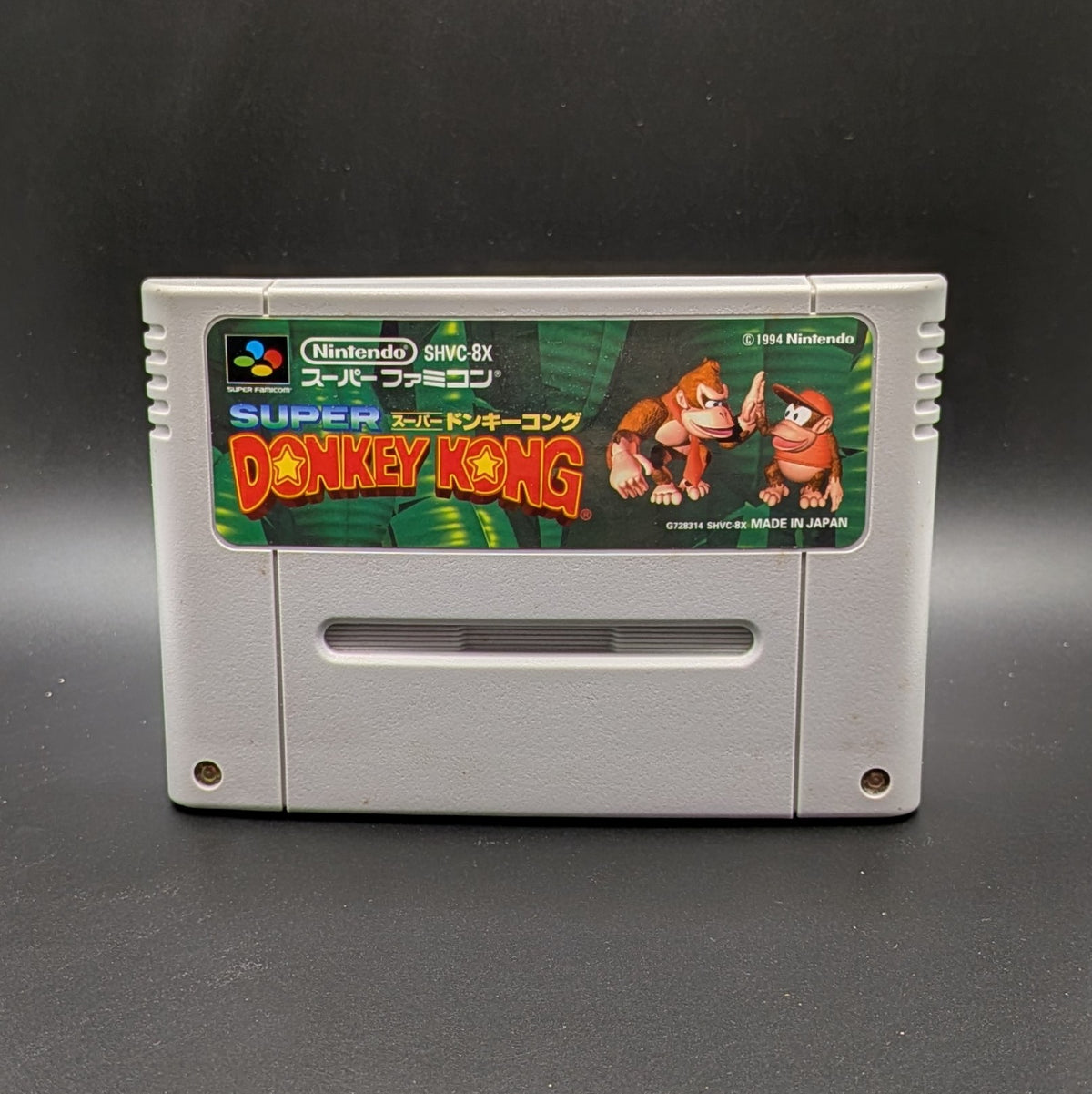 Super Donkey Kong SHVC-8X video game cartridge for the Super Famicom Entertainment System, shown standing up and facing forward on a dark gray background.