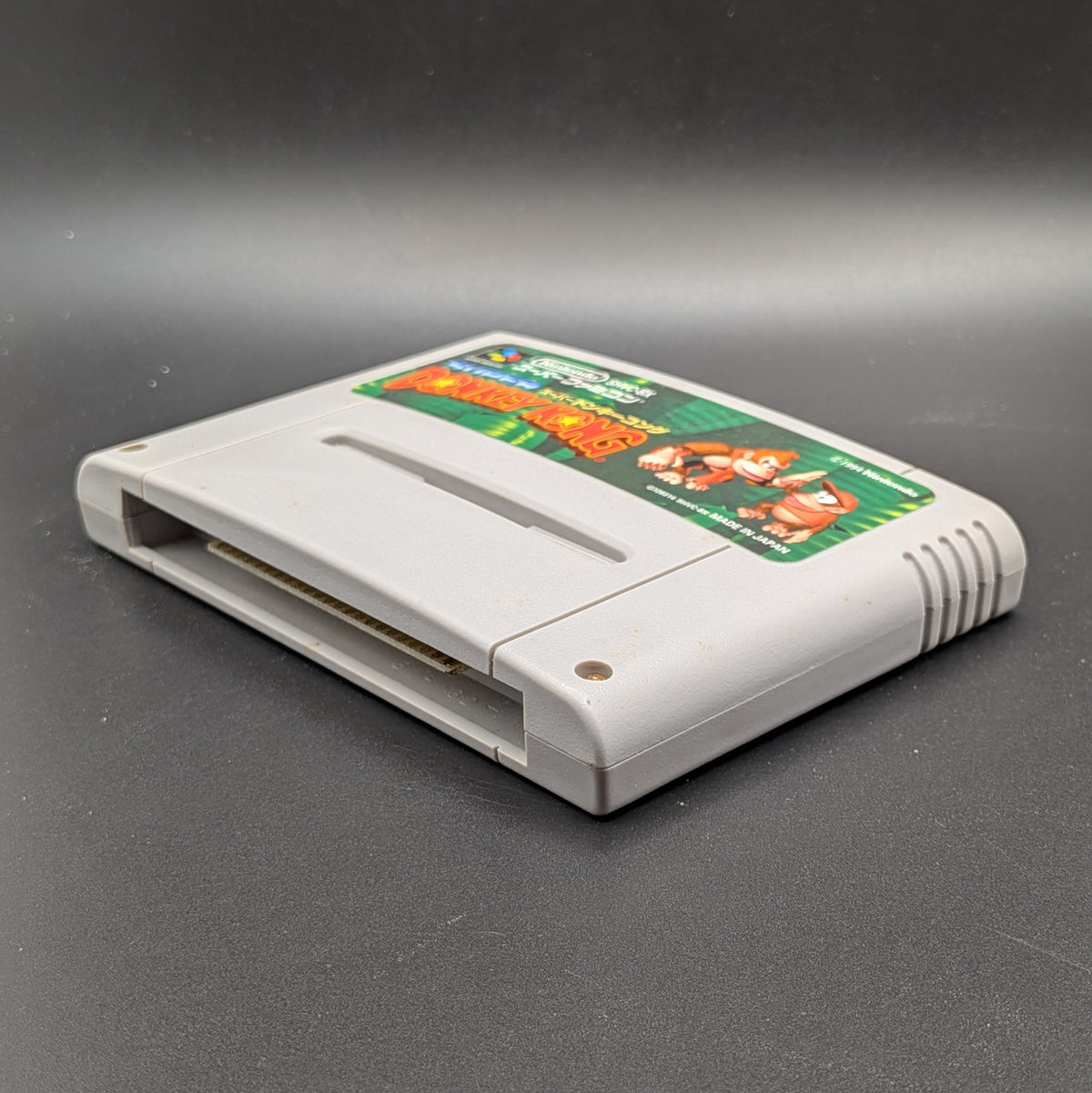 Super Donkey Kong SHVC-8X video game cartridge for the Super Famicom Entertainment System, shown laying on its back, at a 3/4 angle facing up so the label is visible. Pictured on a dark grey background.