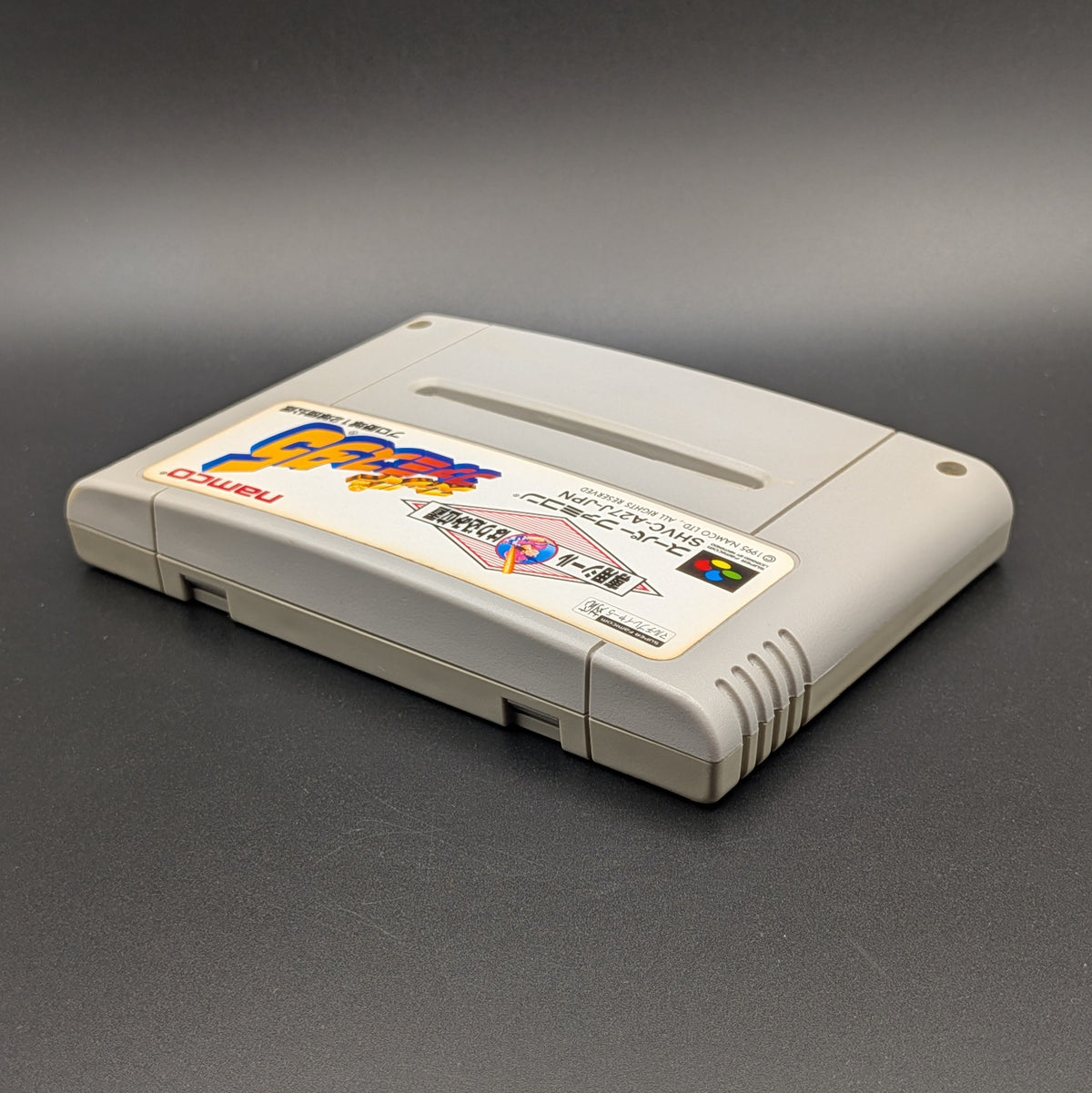 Super Famista 5 video game cartridge for Nintendo's Super Famicom (SFC) Entertainment System, shown laying on its back with the label facing upwards. Showing the left hand side and top edge. It is displayed on a dark grey background.