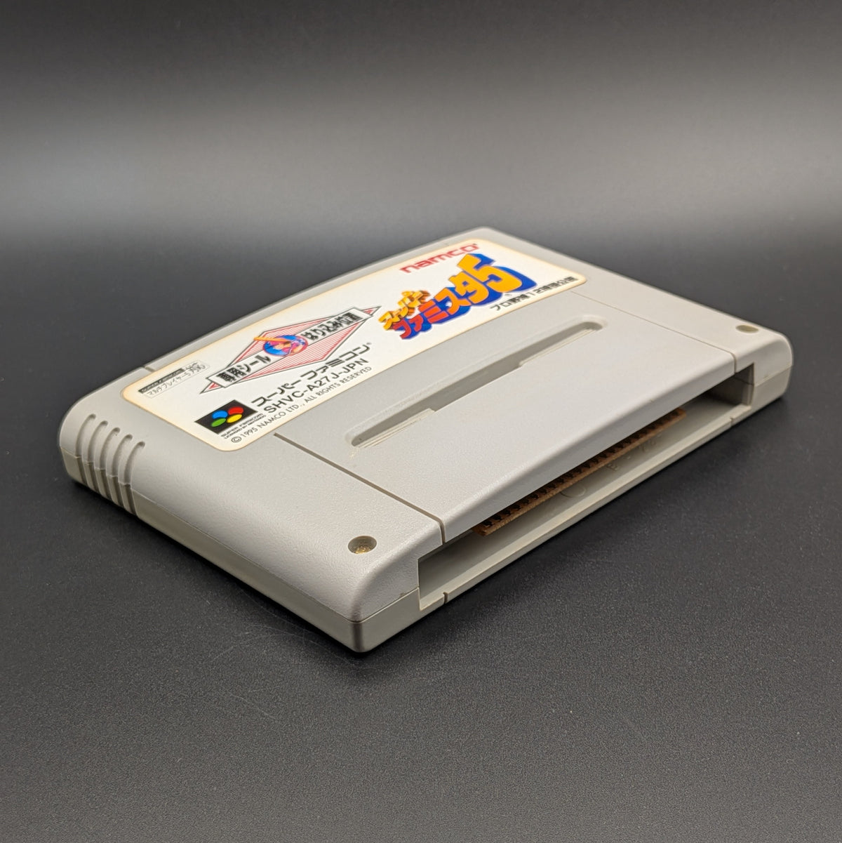 Super Famista 5 video game cartridge for Nintendo's Super Famicom (SFC) Entertainment System, shown laying on its back with the label facing upwards. Showing the left hand side and bottom slot. It is displayed on a dark grey background.