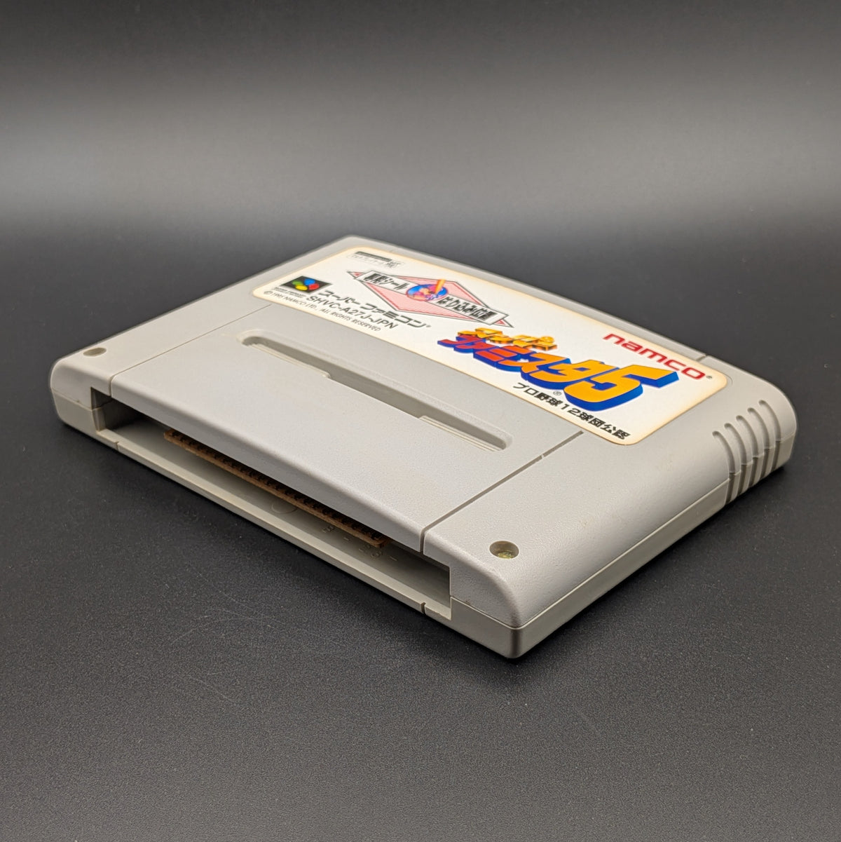 Super Famista 5 video game cartridge for Nintendo's Super Famicom (SFC) Entertainment System, shown laying on its back with the label facing upwards. Showing the right hand side and bottom slot. It is displayed on a dark grey background. 
