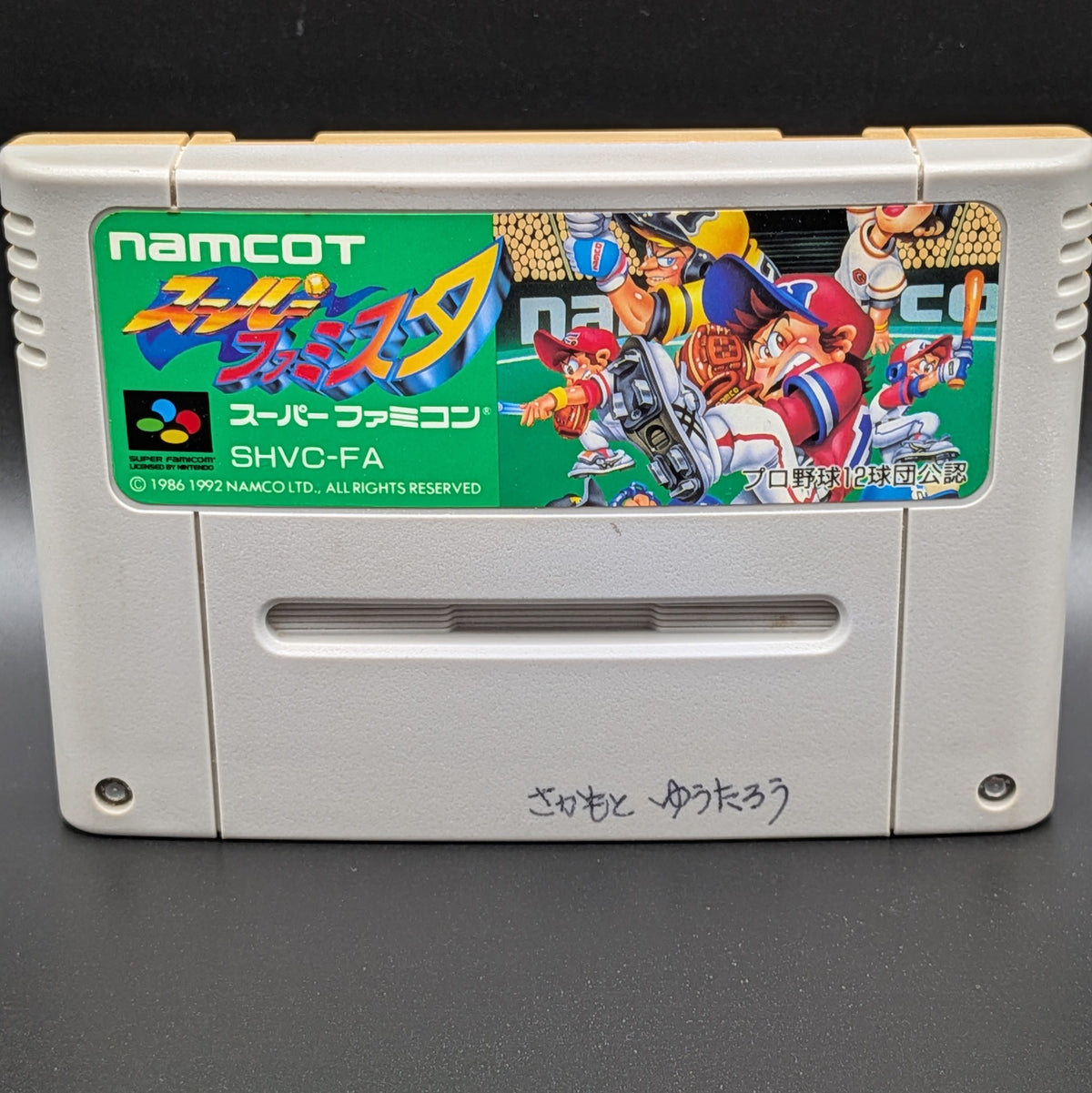 A close up of Super Famista video game cartridge for Nintendo's Super Famicom (SFC) Entertainment System, shown standing up and facing forwards on a dark grey background.
