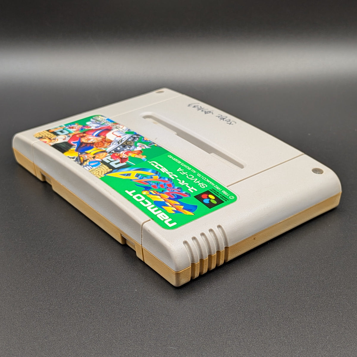 Super Famista video game cartridge for Nintendo's Super Famicom (SFC) Entertainment System, shown laying on its back with the label facing upwards. Showing the left hand side and top edge. It is displayed on a dark grey background.